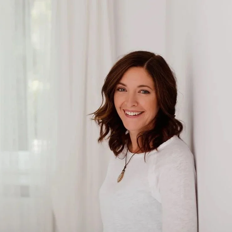 Jasmin Nerici, mit braunen, gewellten Haaren in einem weißen Oberteil vor einem weißen Vorhang in einem Raum mit natürlichem Licht.