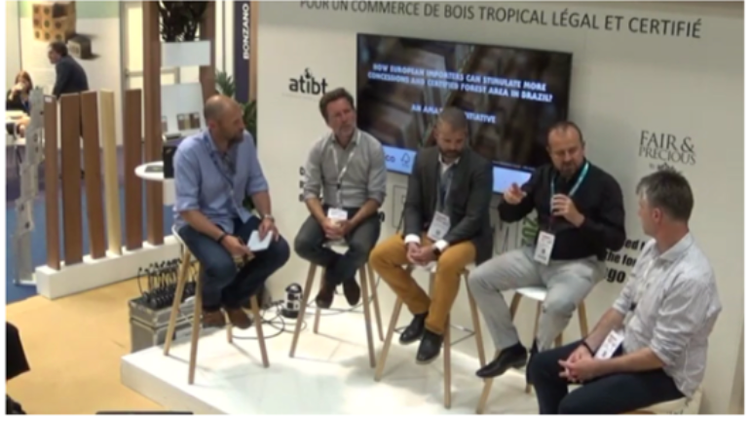 Five men seated on stools participating in a panel discussion at a trade show or conference, with a large screen and branding in the background.