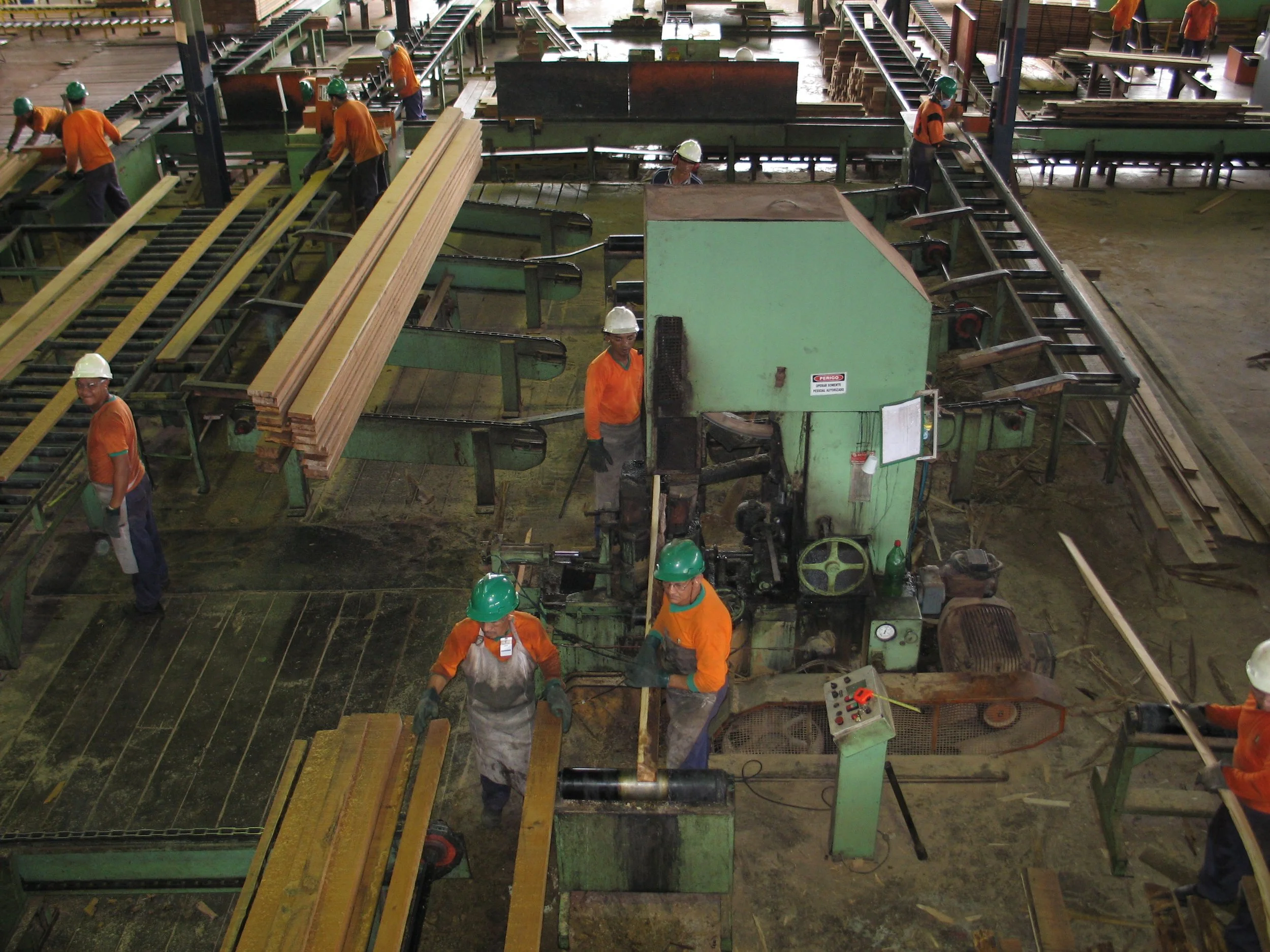 Workers in an industrial plant operating machinery for processing wood, with some handling large wooden planks and others working at conveyor belts.