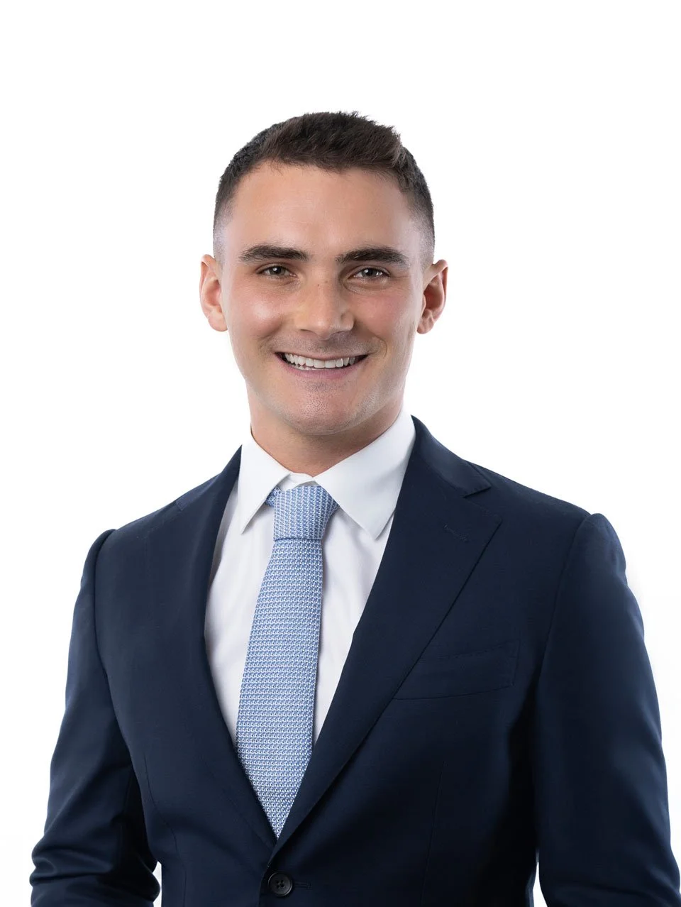 A young man with short dark hair, wearing a navy suit, white shirt, and light blue tie, smiling against a white background.