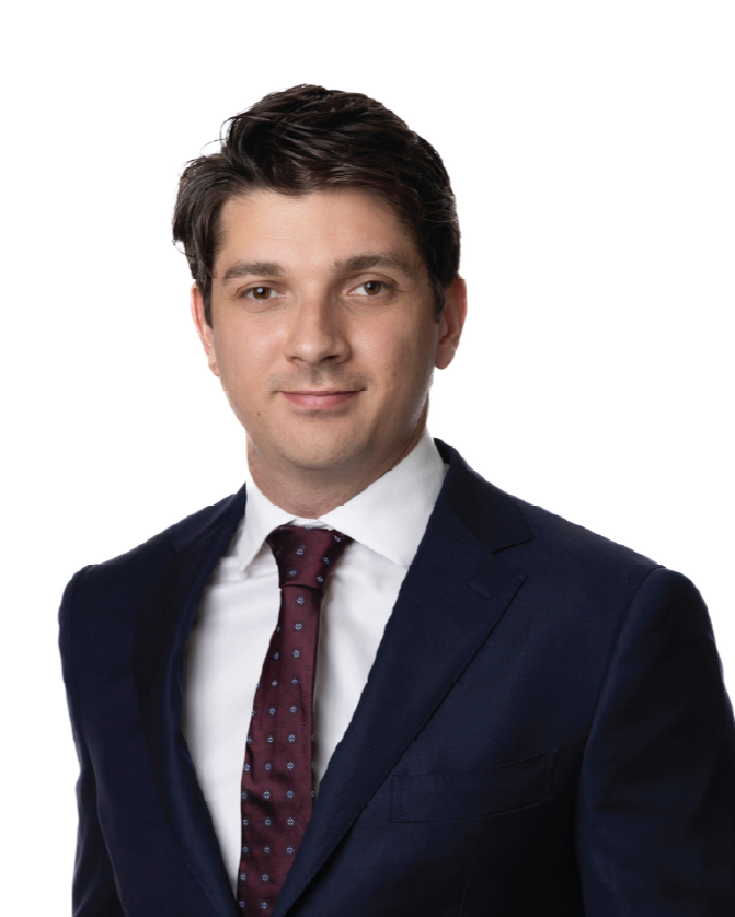 A man with dark brown hair, wearing a dark suit, white shirt, and maroon tie, smiling slightly against a plain white background.