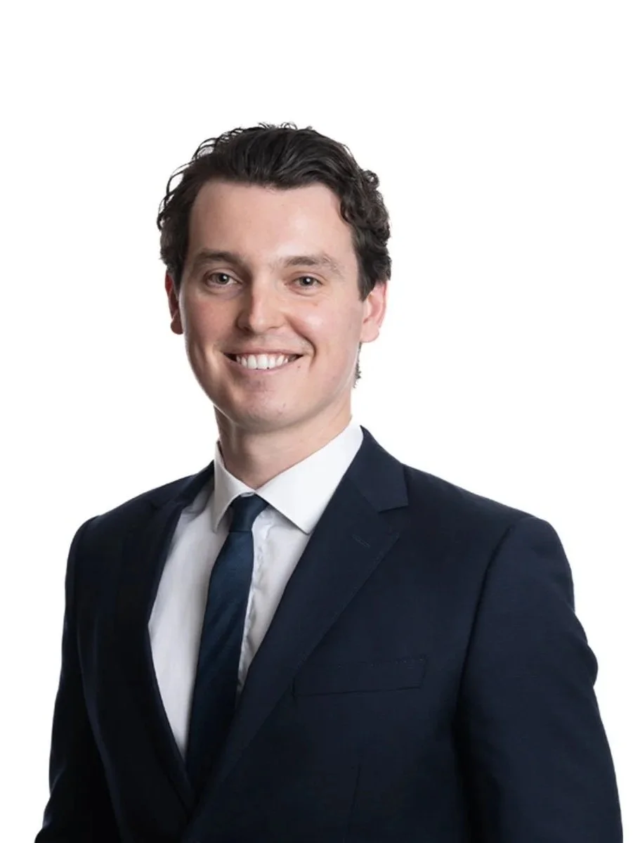 A young man with dark, curly hair wearing a dark suit, white shirt, and dark tie against a white background, smiling at the camera.