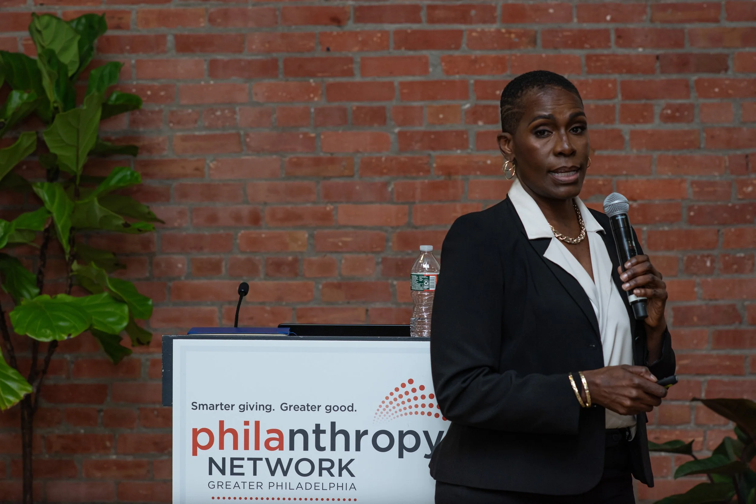 Woman giving a presentation at a philanthropy event, holding a microphone, with a sign that reads 'Smarter giving. Greater good. philanthropy NETWORK GREATER PHILADELPHIA' in front of a red brick wall.