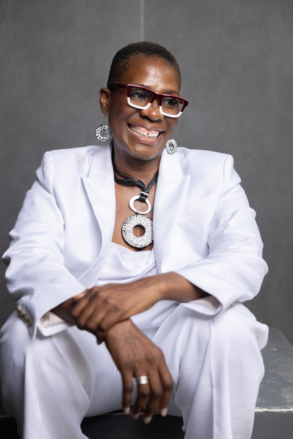 A smiling woman with short hair, wearing red and white glasses, earrings, and a white outfit with jewelry, sitting against a neutral background.