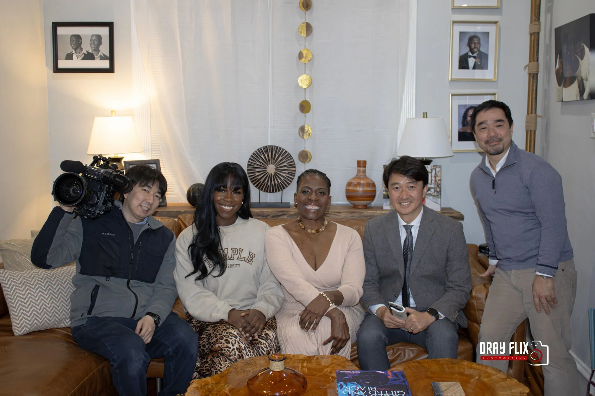Group of five people sitting on a leather sofa in a living room, smiling at the camera, with a cameraman standing on the left holding a video camera. The background features framed photos on the wall, lamps, vases, and decorative items on a side table.