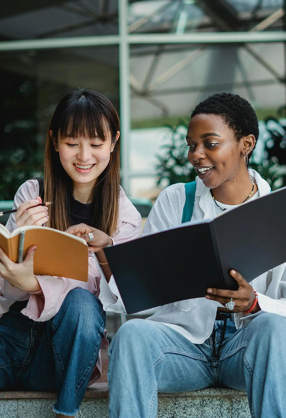 Two women sitting outdoors, looking at a notebook and smiling, one holding a pen and the other holding a folder, in a casual setting with greenery in the background.