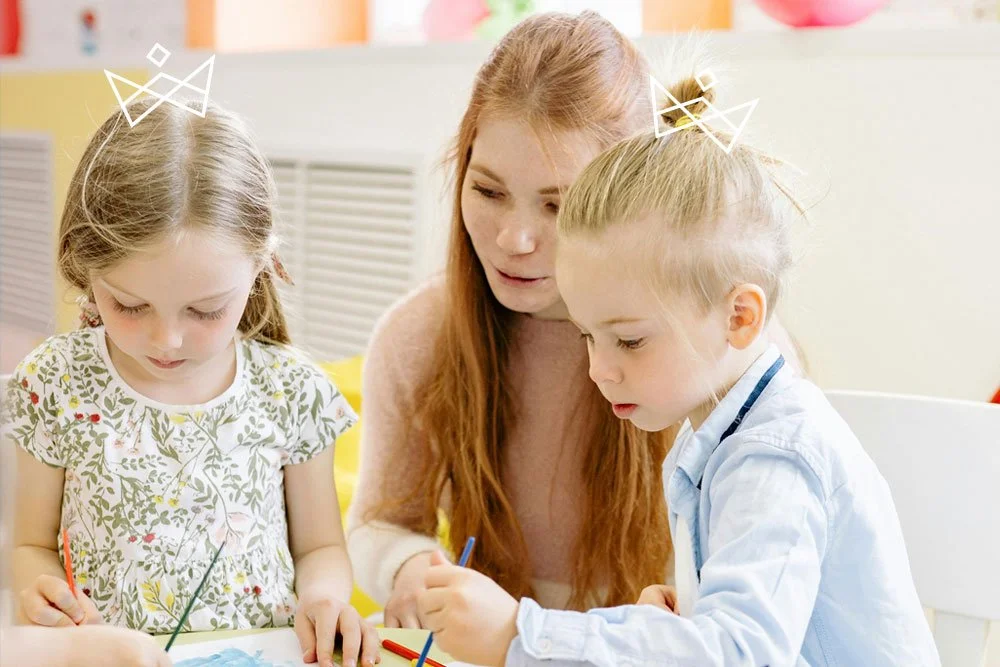 A woman teaching two young girls how to paint at a table in a colorful classroom.