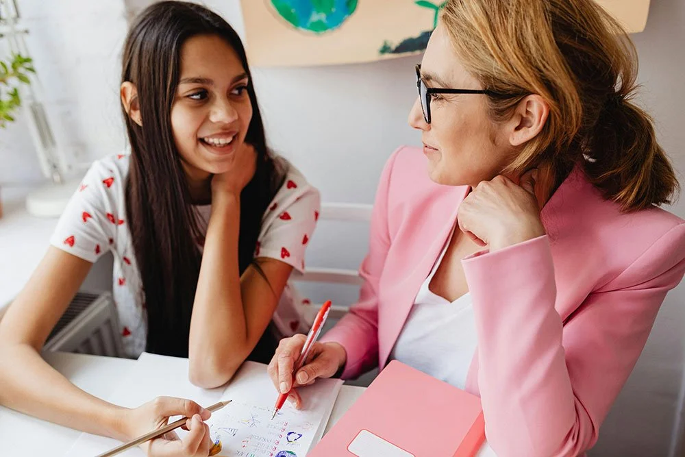 A young girl and an adult woman are sitting at a table, smiling and talking to each other. The girl has long dark hair and wears a white shirt with red hearts. The woman has blonde hair, glasses, and is wearing a pink blazer. There are notebooks, pens, and a colorful drawing on the table.