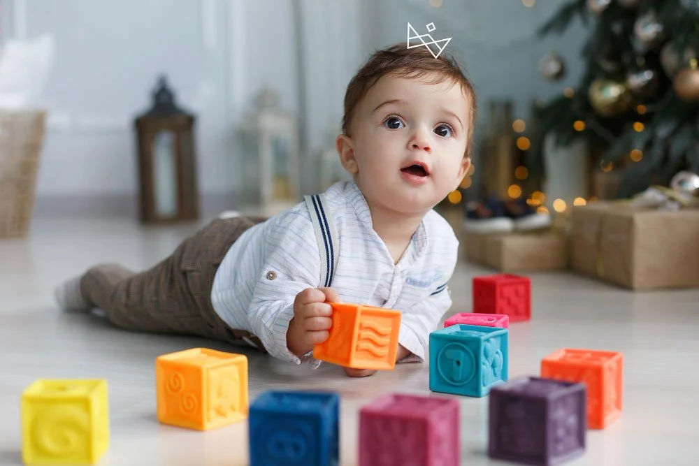 A young boy lies on the floor surrounded by colorful toy blocks, holding an orange block, with a decorated Christmas tree and wrapped presents in the background.