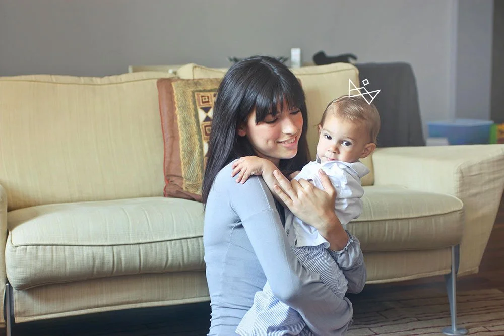 A woman with black hair smiling while holding a baby in a living room with a beige couch and colorful cushions.