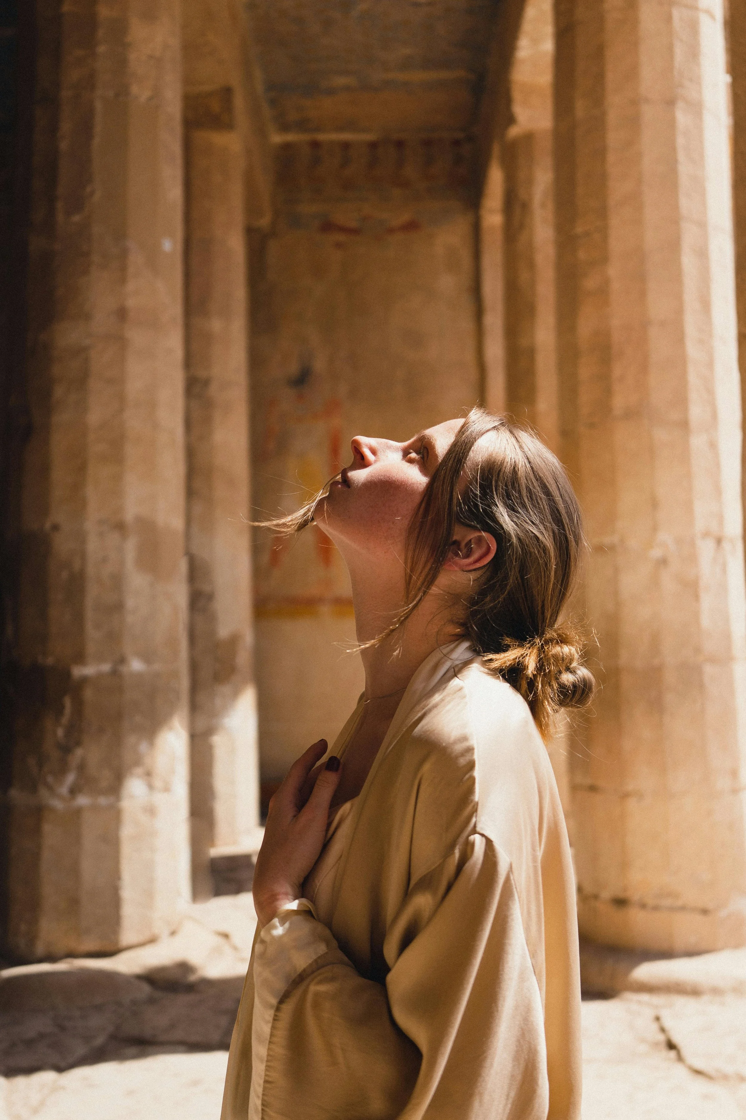 A woman in beige clothing standing in an ancient stone structure, looking upwards with her eyes closed, one hand touching her chest.