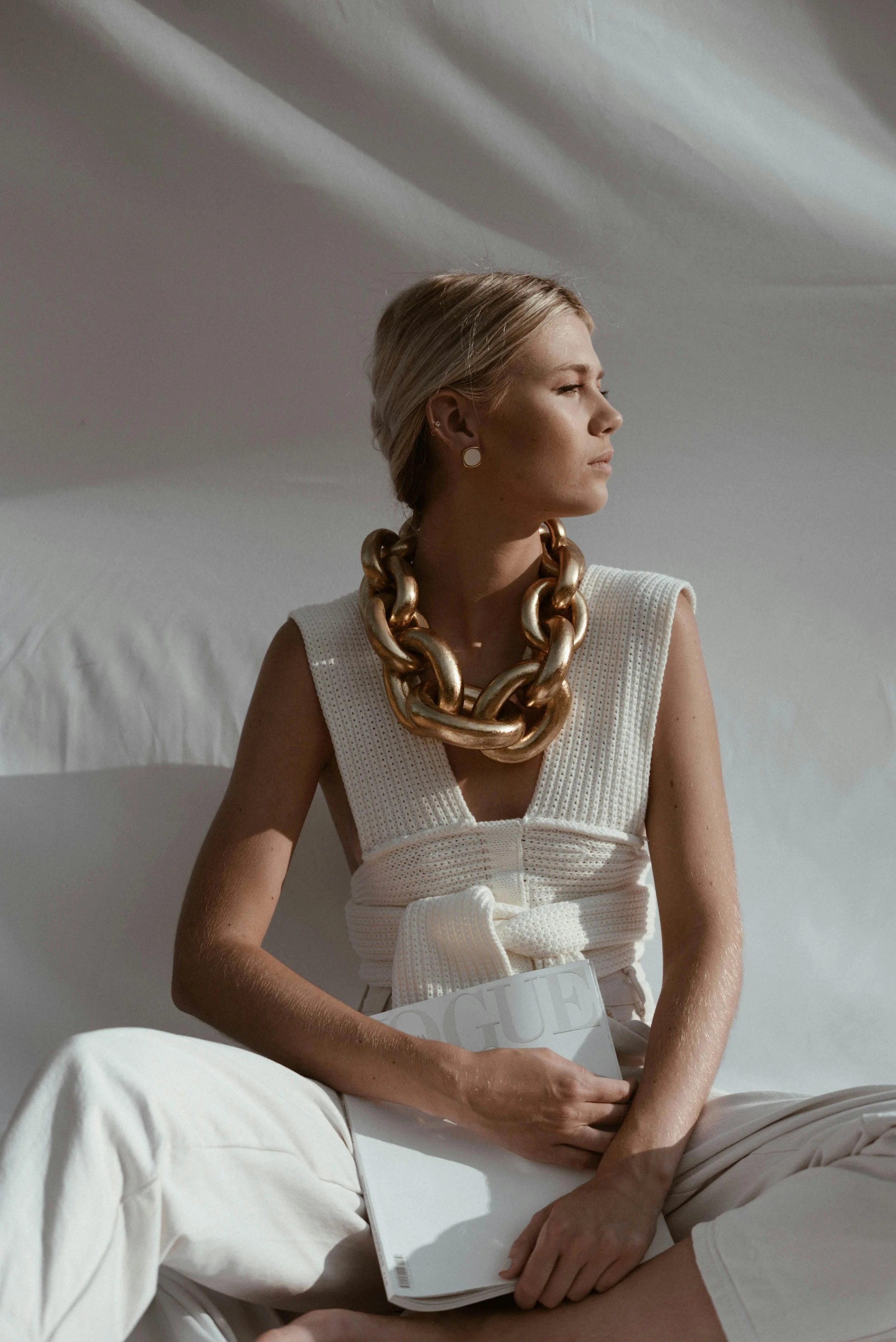 Woman sitting on bed, wearing a white sleeveless top, chunky gold necklace, and earrings, holding a Vogue magazine, with beige pants and plain background.