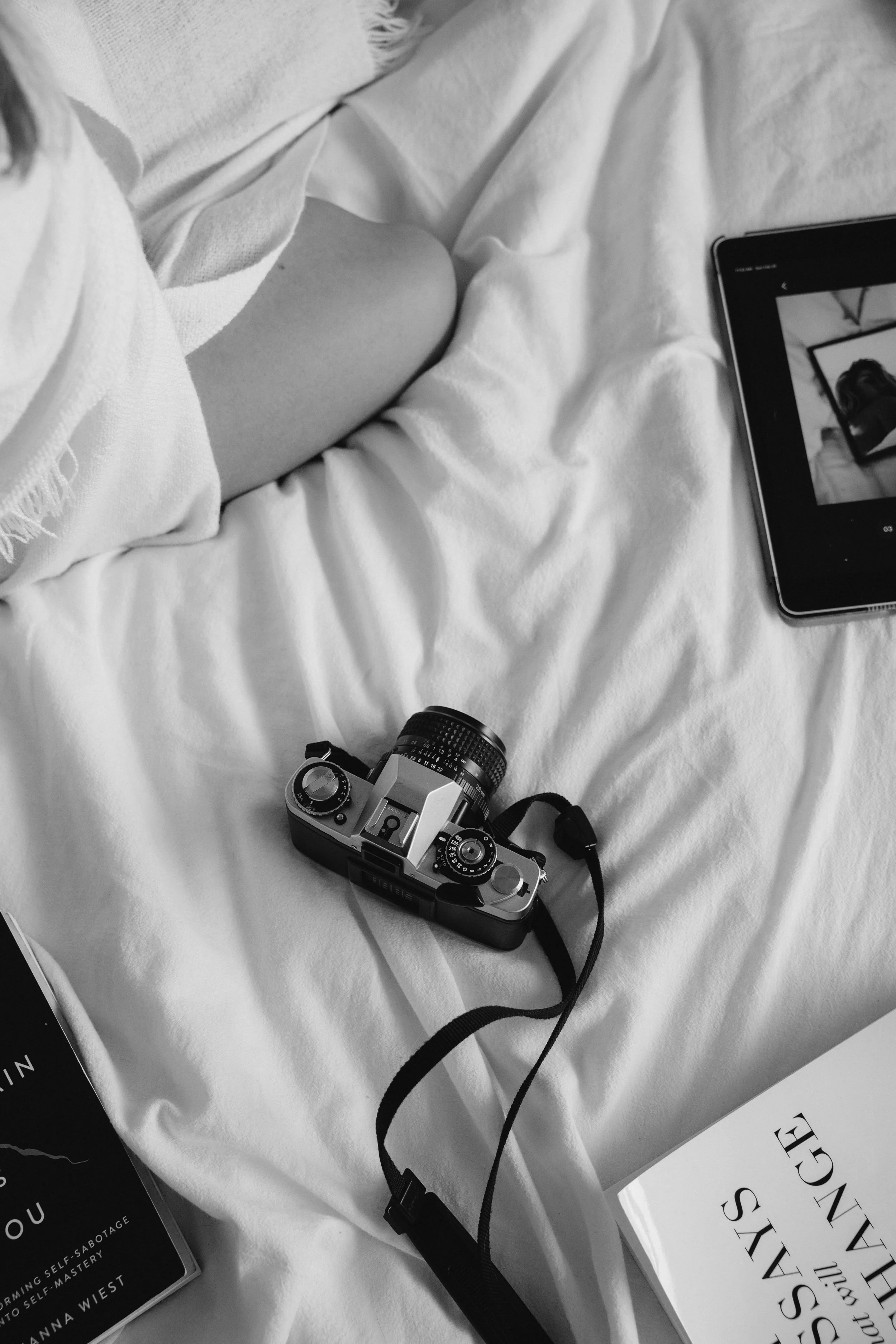 A black and white photo showing a bed with a camera, a tablet, a book, and part of a person’s leg and white shirt.