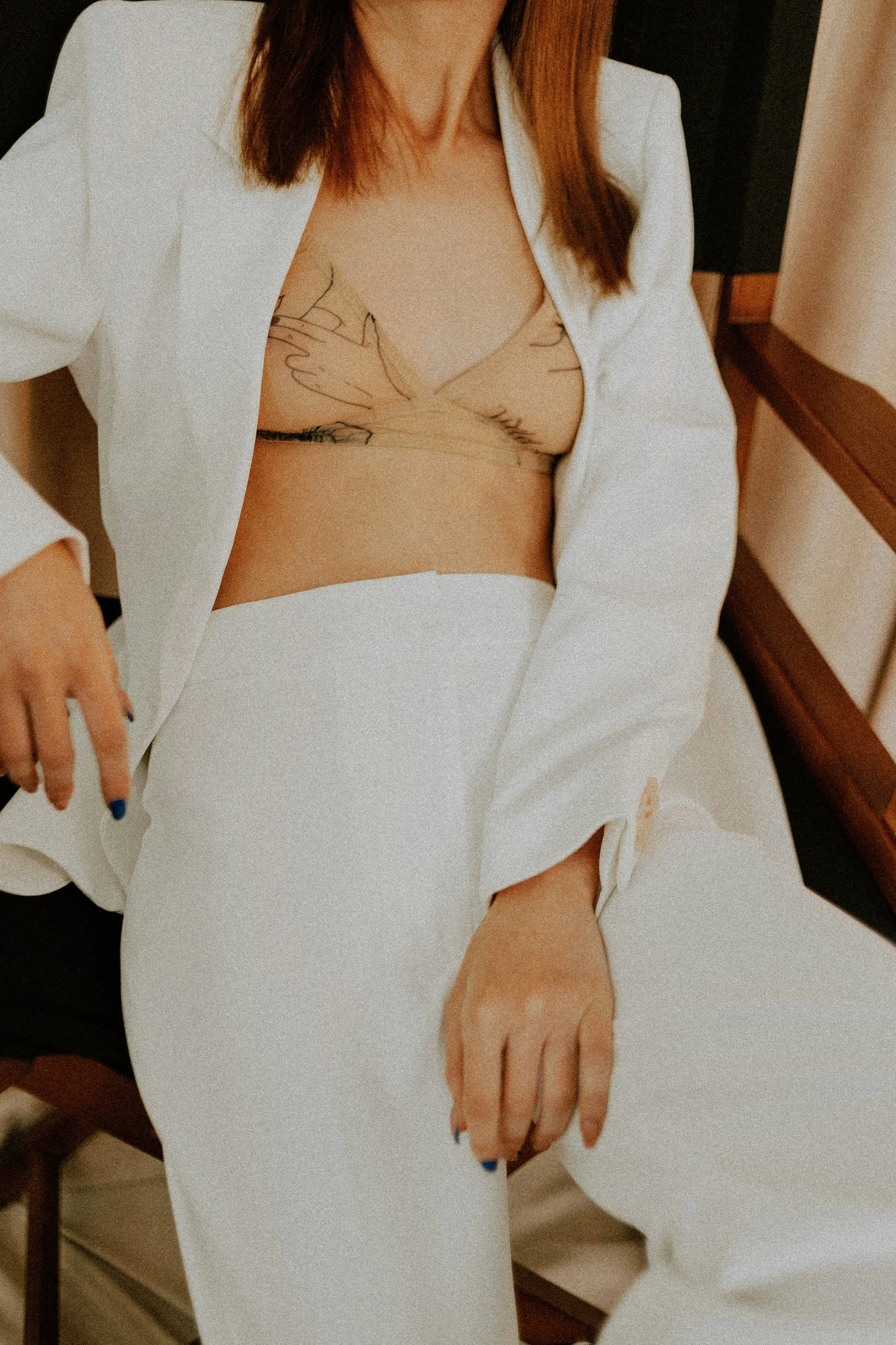 A woman sitting on a chair, wearing a white blazer and white pants, with a sheer beige top that has black line art tattoos on her chest.