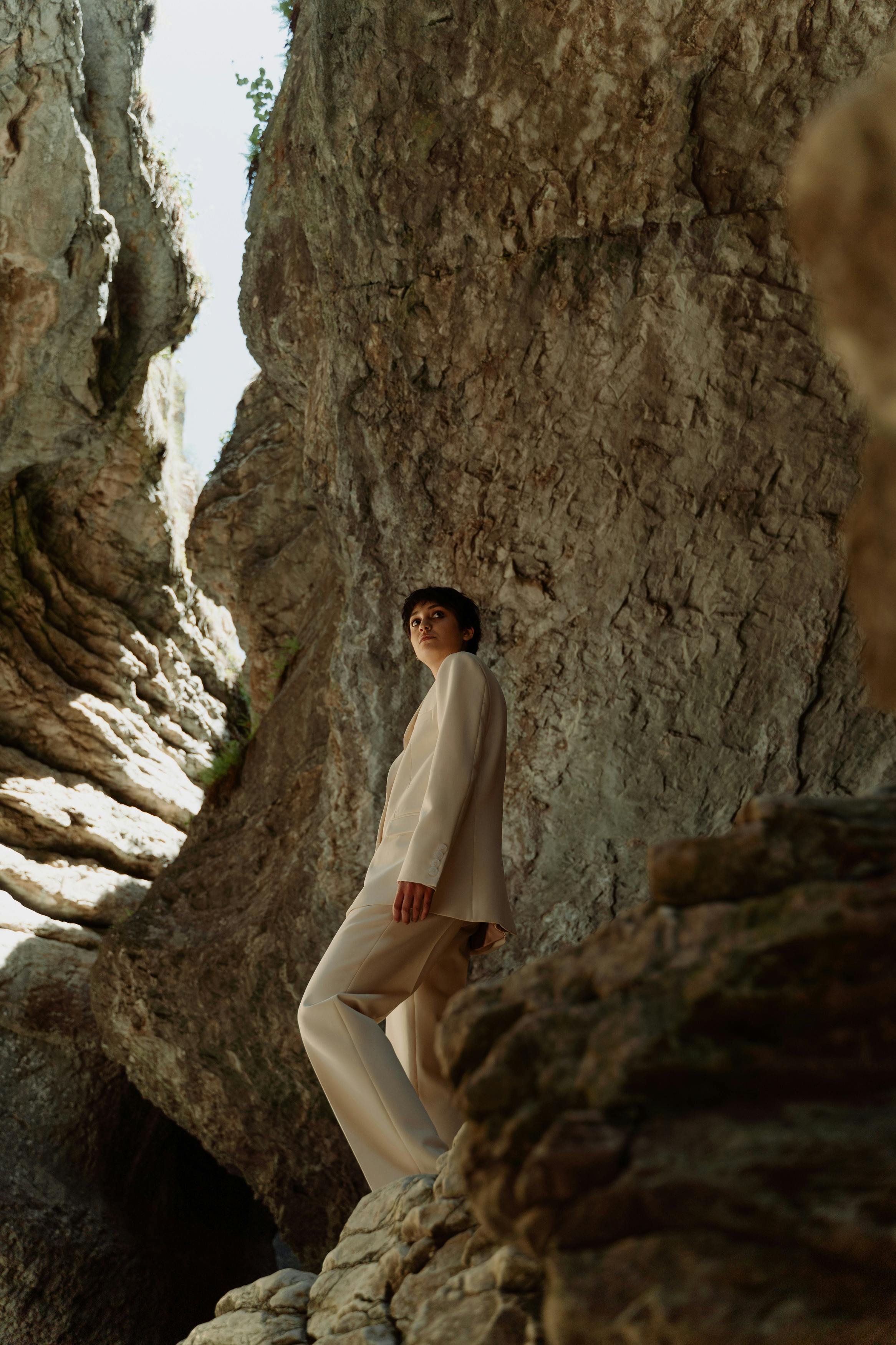 A woman in a beige pantsuit standing amidst large rocks and cliffs, with a rocky crevice and sky visible in the background.