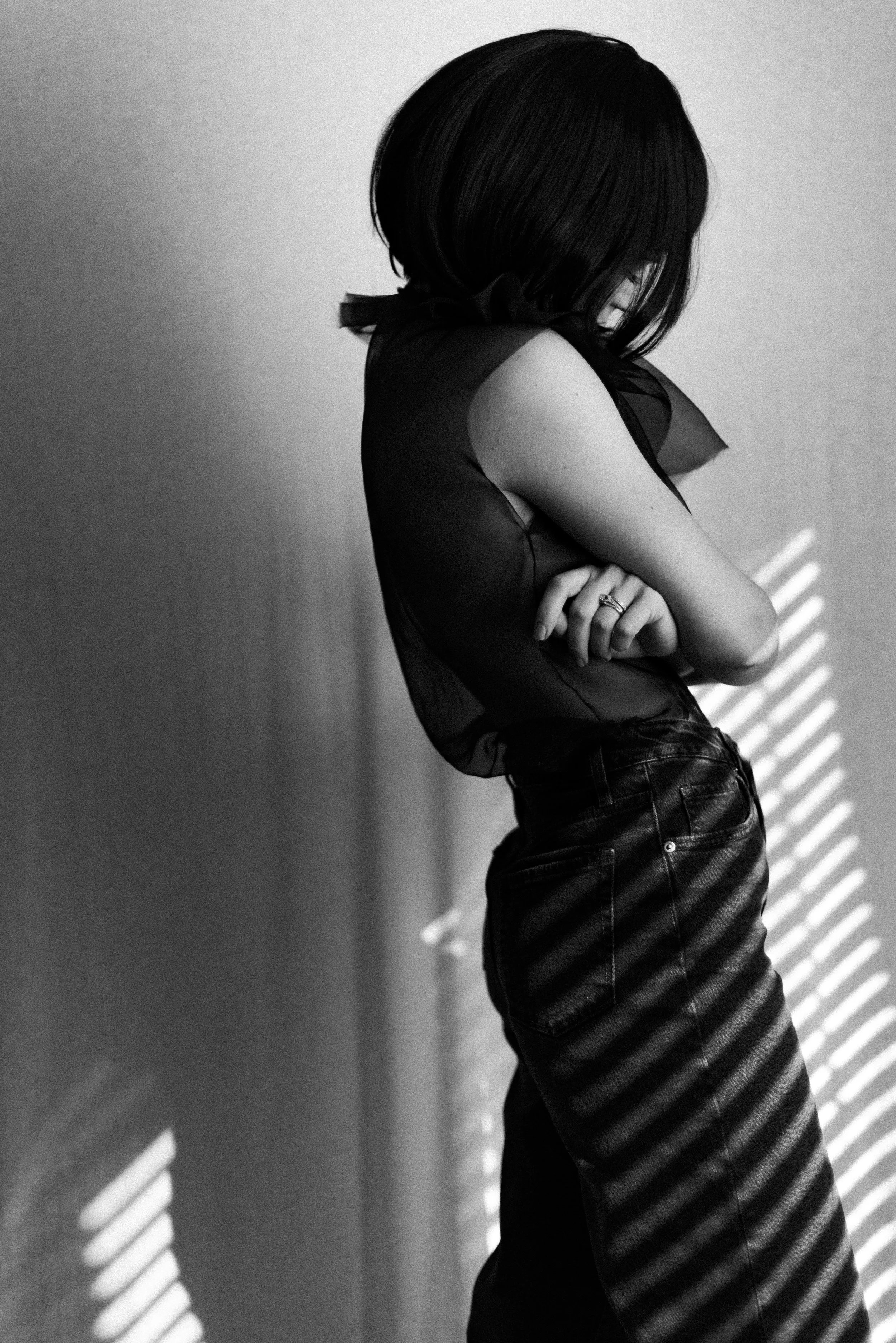 A woman with shoulder-length dark hair stands against a plain wall, with shadows of window blinds cast on her. She is wearing a sleeveless top with a bow at the neck and high-waisted pants, with her arms crossed.