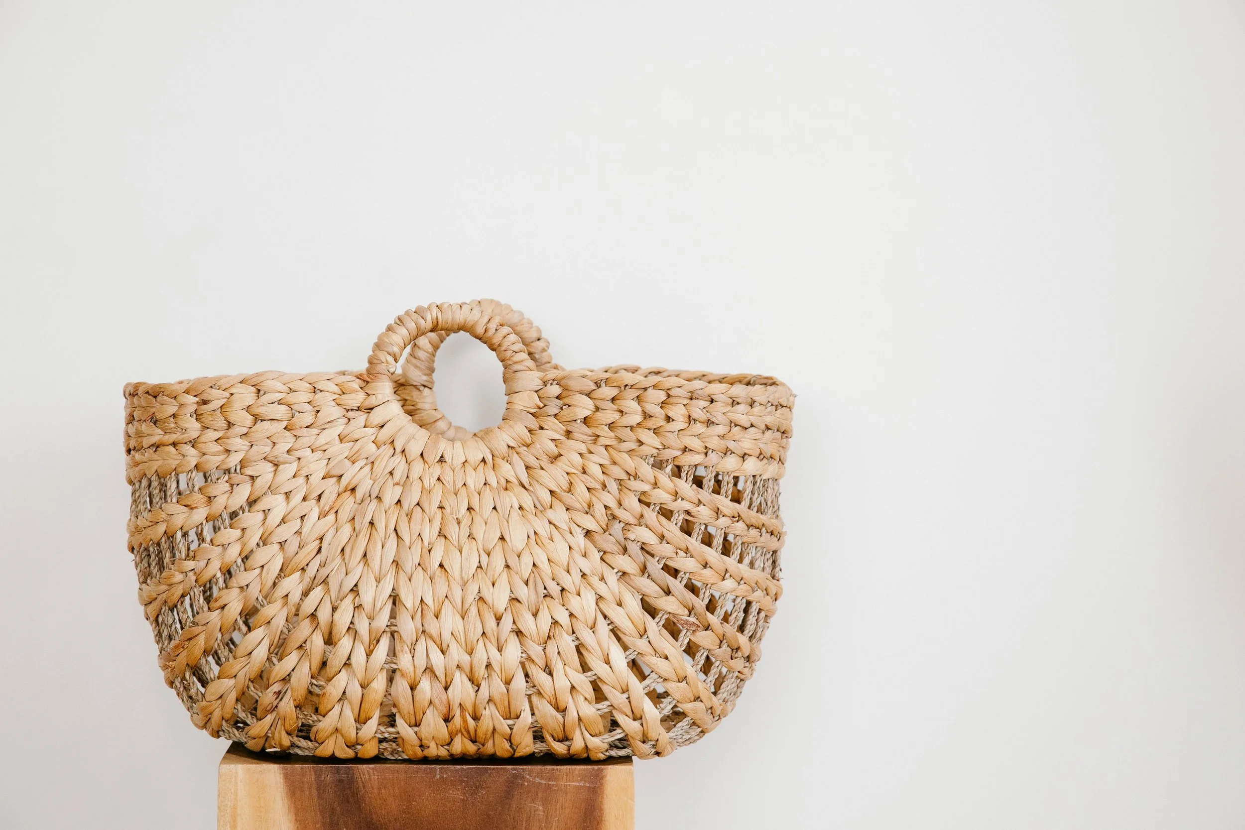 A woven straw handbag with round handles resting on a wooden surface against a plain white background.