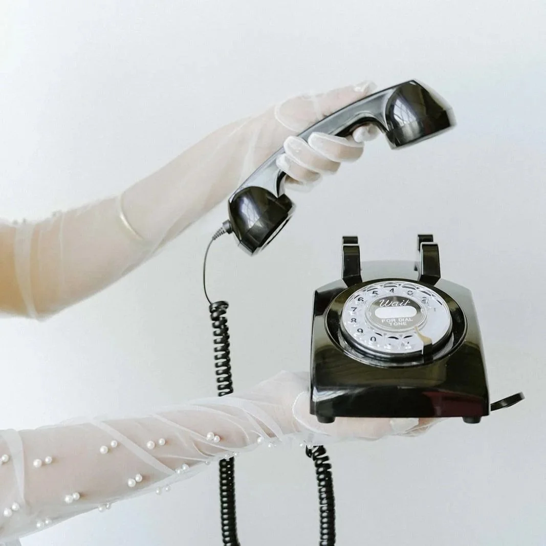 Person wearing white gloves holding a black rotary phone with a cord.