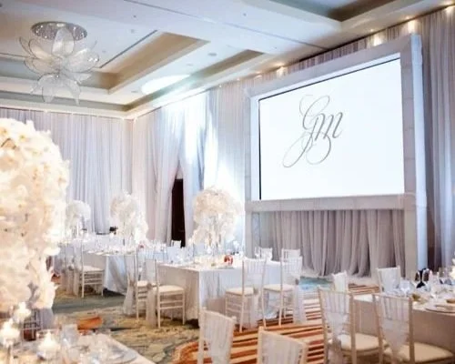 Elegant event space with round tables, white chairs, floral centerpieces, and a large screen displaying initials 'G M', decorated in soft neutral and white tones.