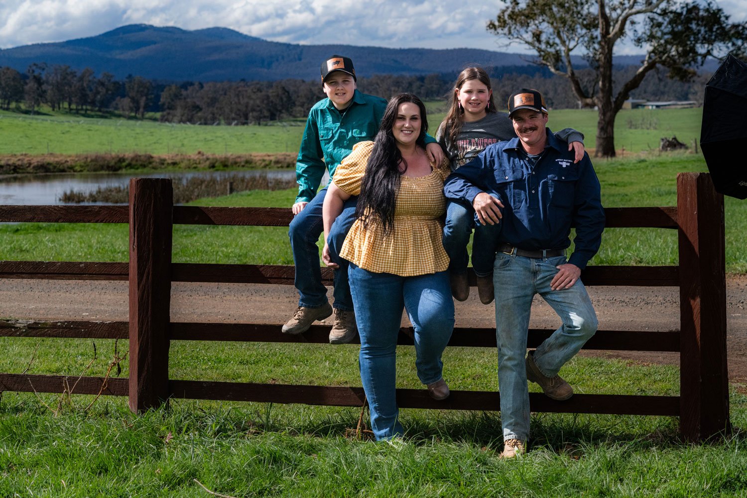 A family of five posing outdoors on a farm with green grass, a pond, and mountains in the background. Two children are sitting on a wooden fence, with a woman standing in front and a man standing beside her.