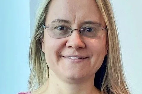 A woman with blonde hair wearing glasses and a pink shirt, smiling at the camera.
