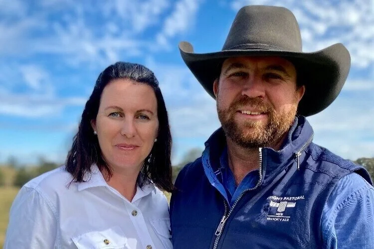 A man and woman standing outdoors under a partly cloudy sky, with the man wearing a cowboy hat and a blue jacket, and the woman wearing a white shirt.