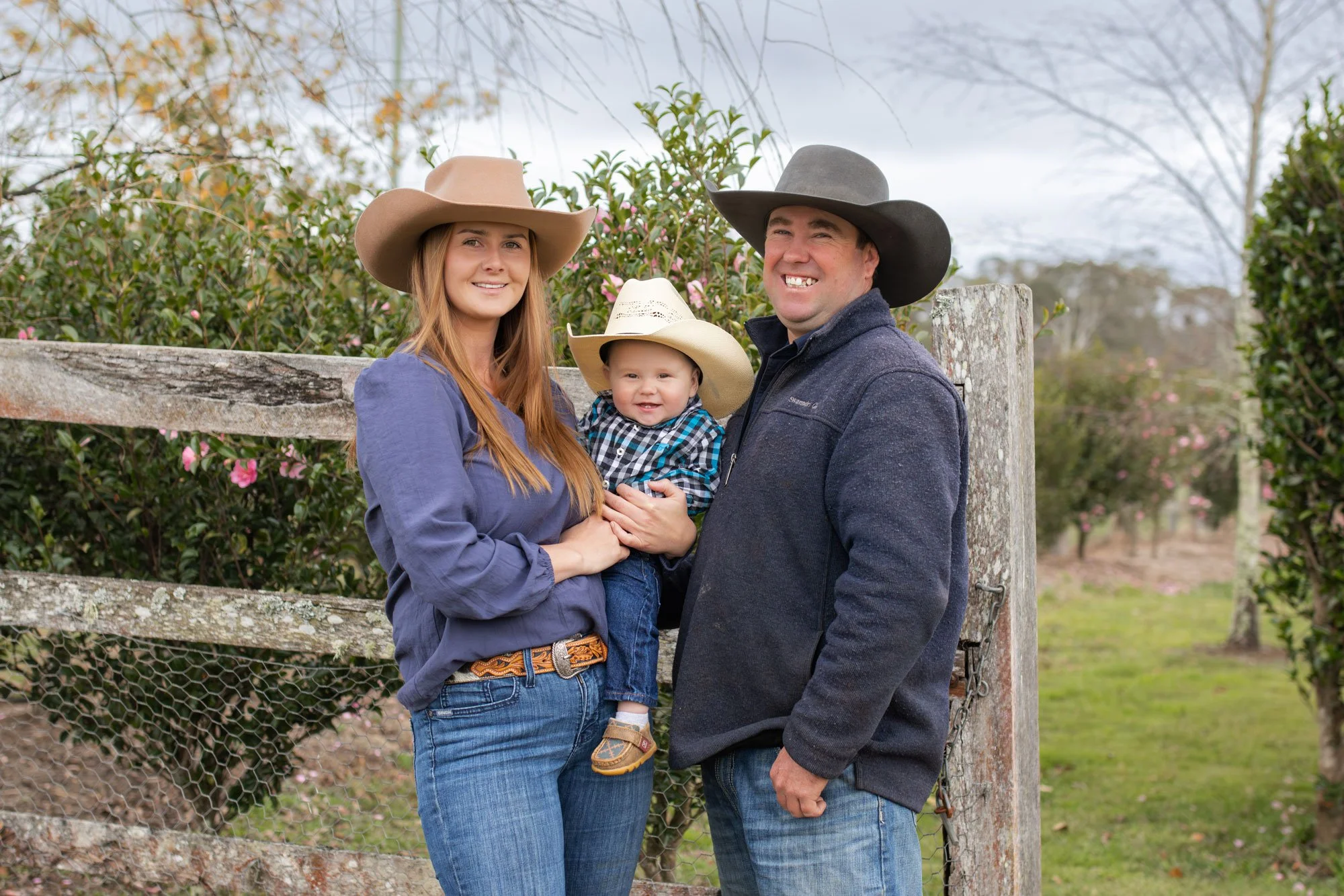 Family photo of a woman, man, and a young child in cowboy hats outdoors near a wooden fence and pink flowering bushes.