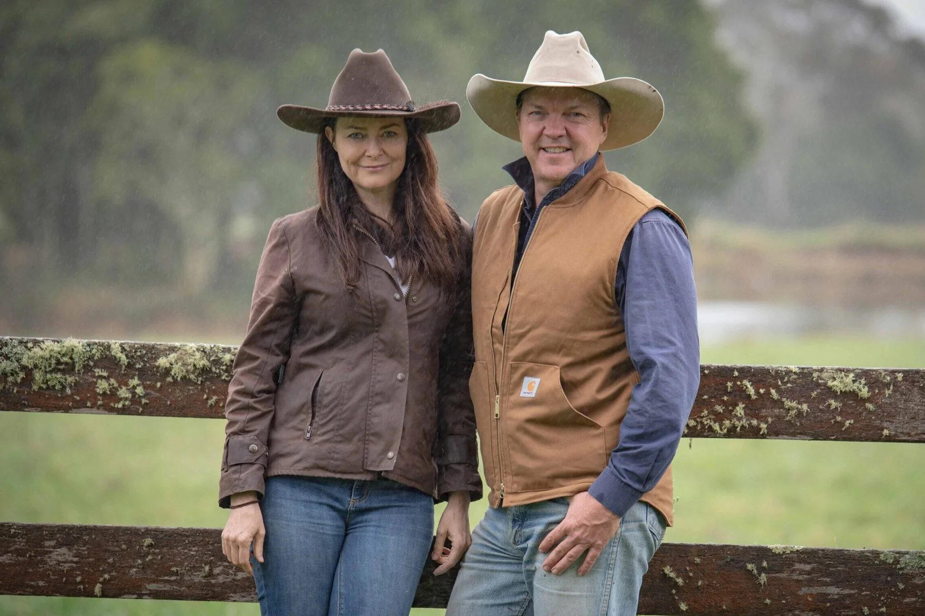 Current custodians Juliette and Glen Wright, standing outdoors in front of a weathered wooden fence with moss, both wearing wide-brimmed hats, during a rainy day with a green pasture behind them.