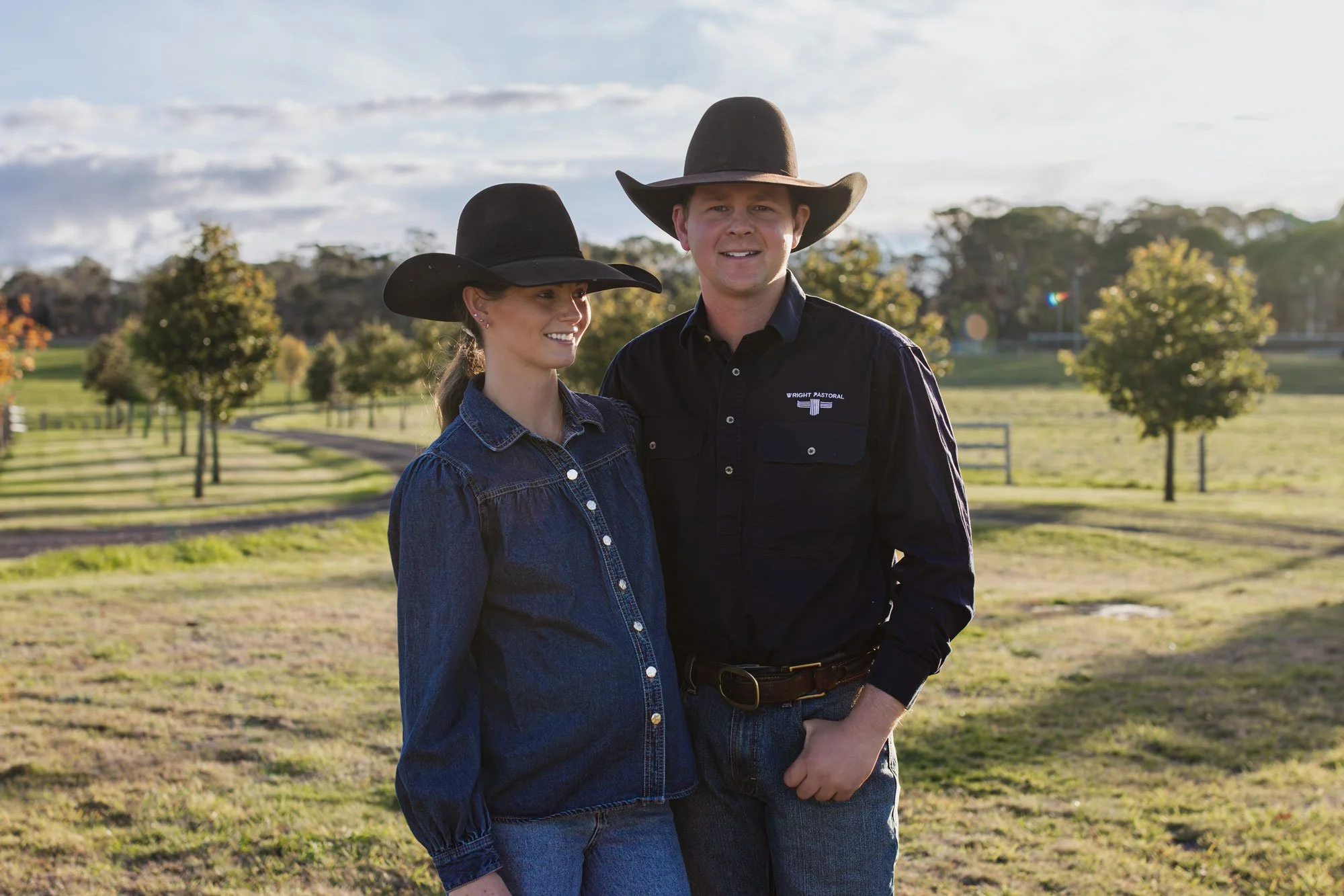 A man and a woman standing outdoors on a farm or ranch, both wearing cowboy hats and denim clothing, smiling and looking at the camera.