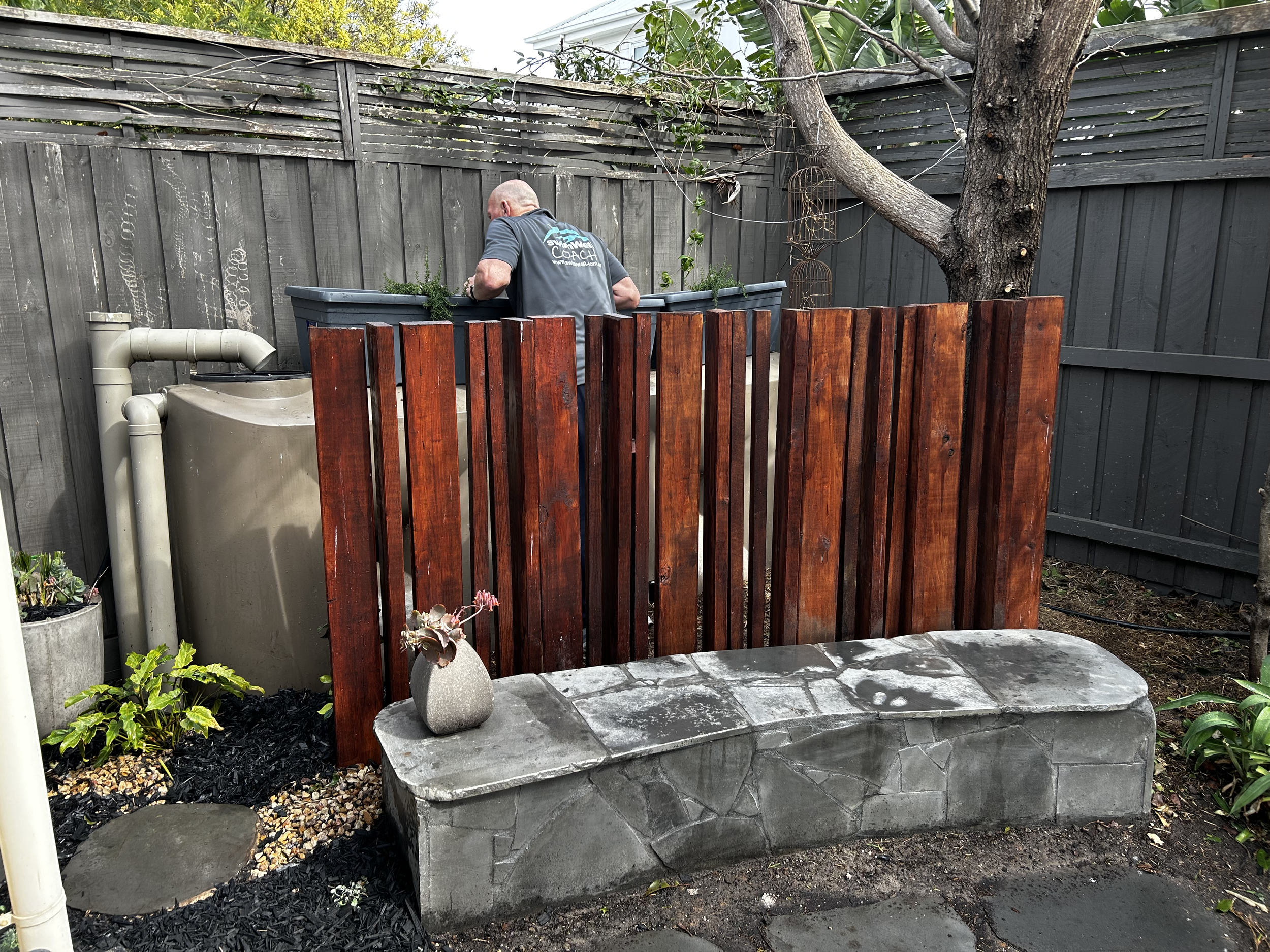 A man with a shaved head, wearing a gray shirt, is working in a backyard garden behind a wooden fence. There is a large grey water collection tank, a concrete bench with a vase of flowers on it, a potted plant, and a tree with a thick trunk in the sc
