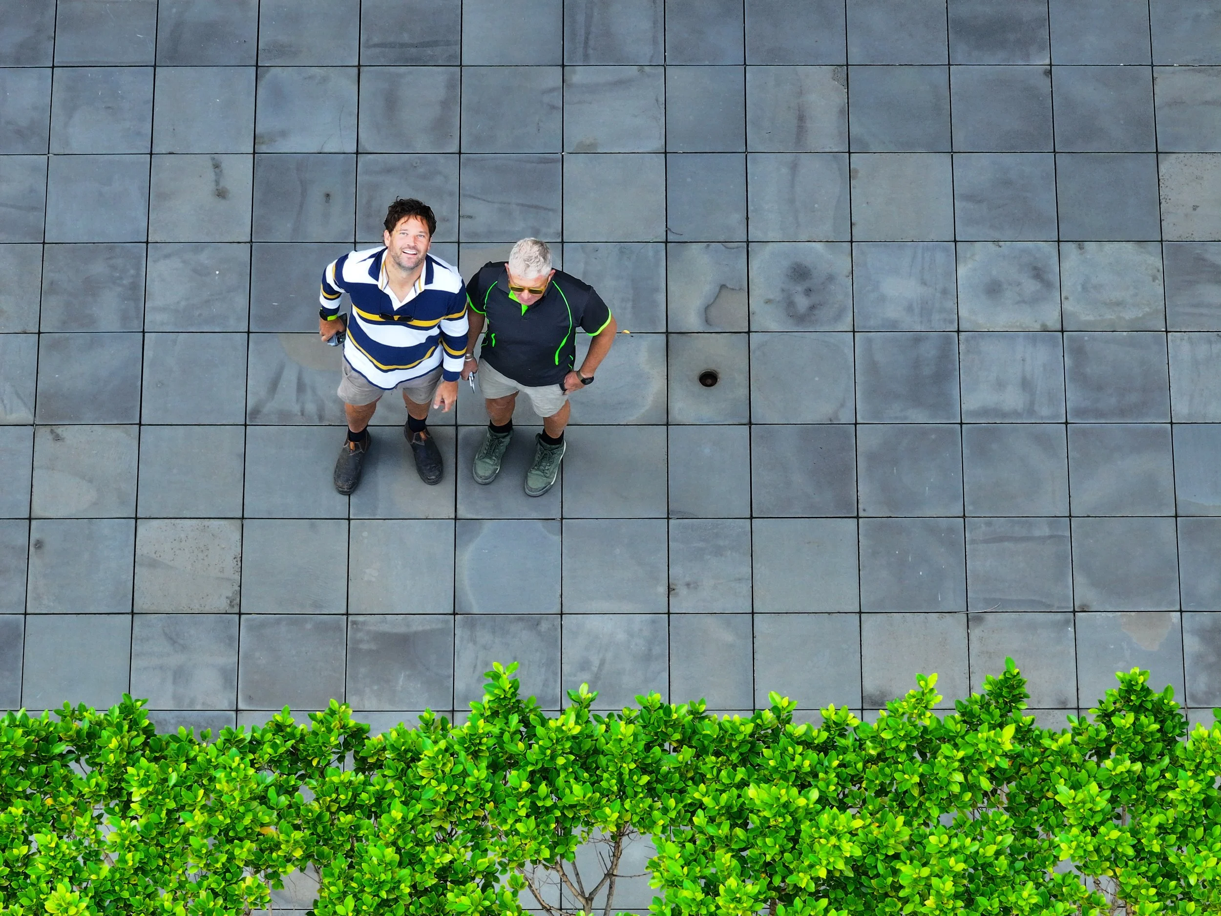 Two men standing on a tiled sidewalk, looking up at the camera, with green bushes in the foreground.