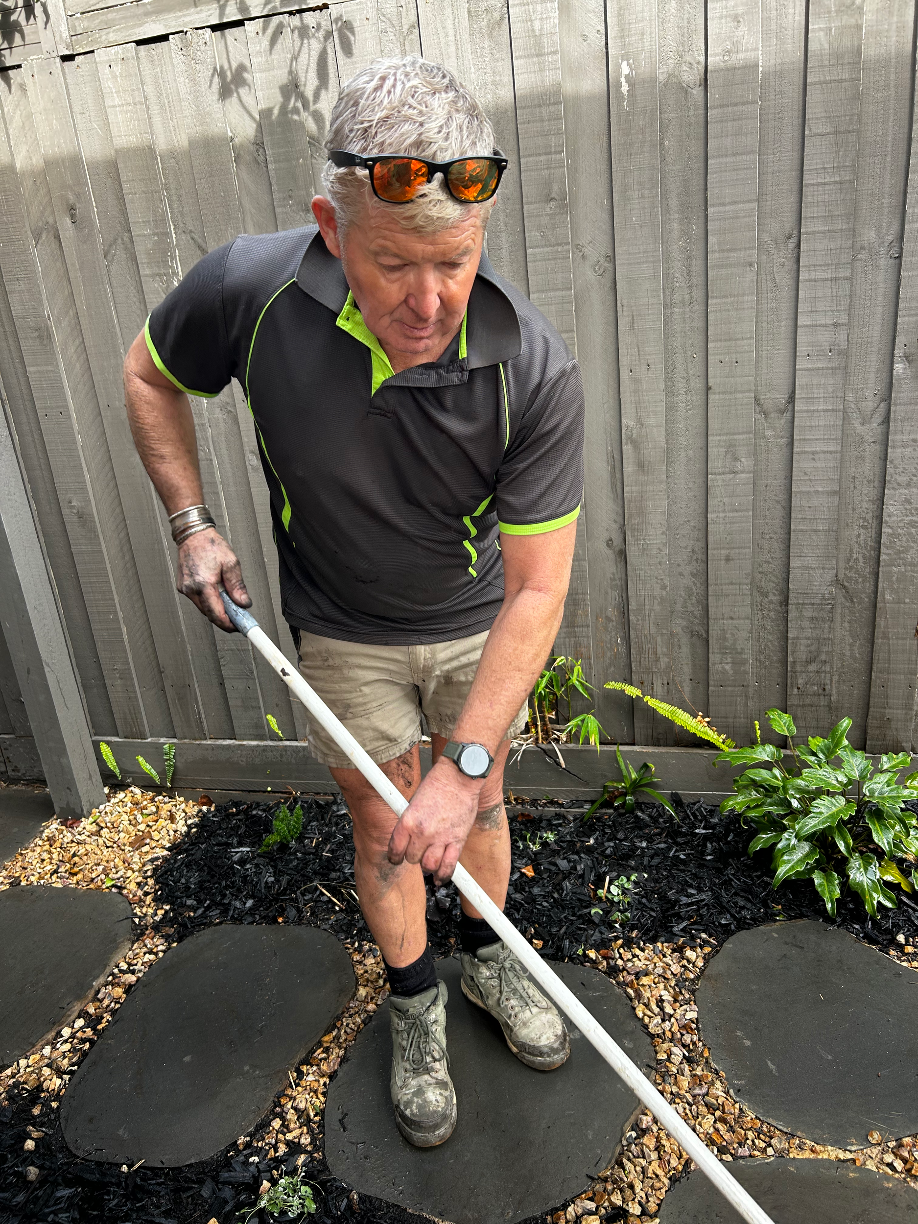 A man planting in a garden, standing on stepping stones with a gardening tool, wearing a black and green shirt, beige shorts, sunglasses on his head, and a watch.
