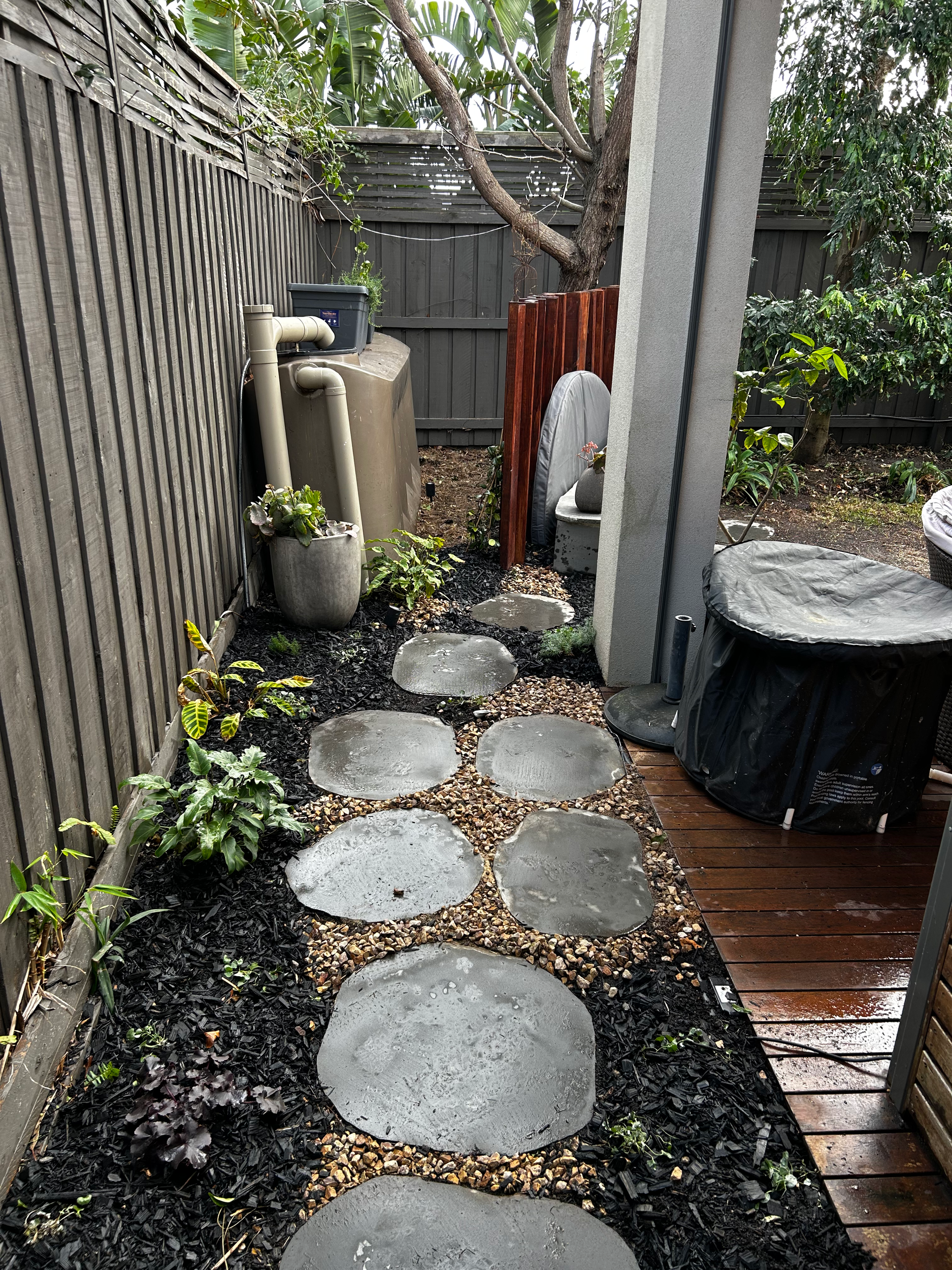 Wet stone pathway in a backyard garden next to wooden deck, surrounded by plants and trees, with a hot tub covered with a black cover on the right side.