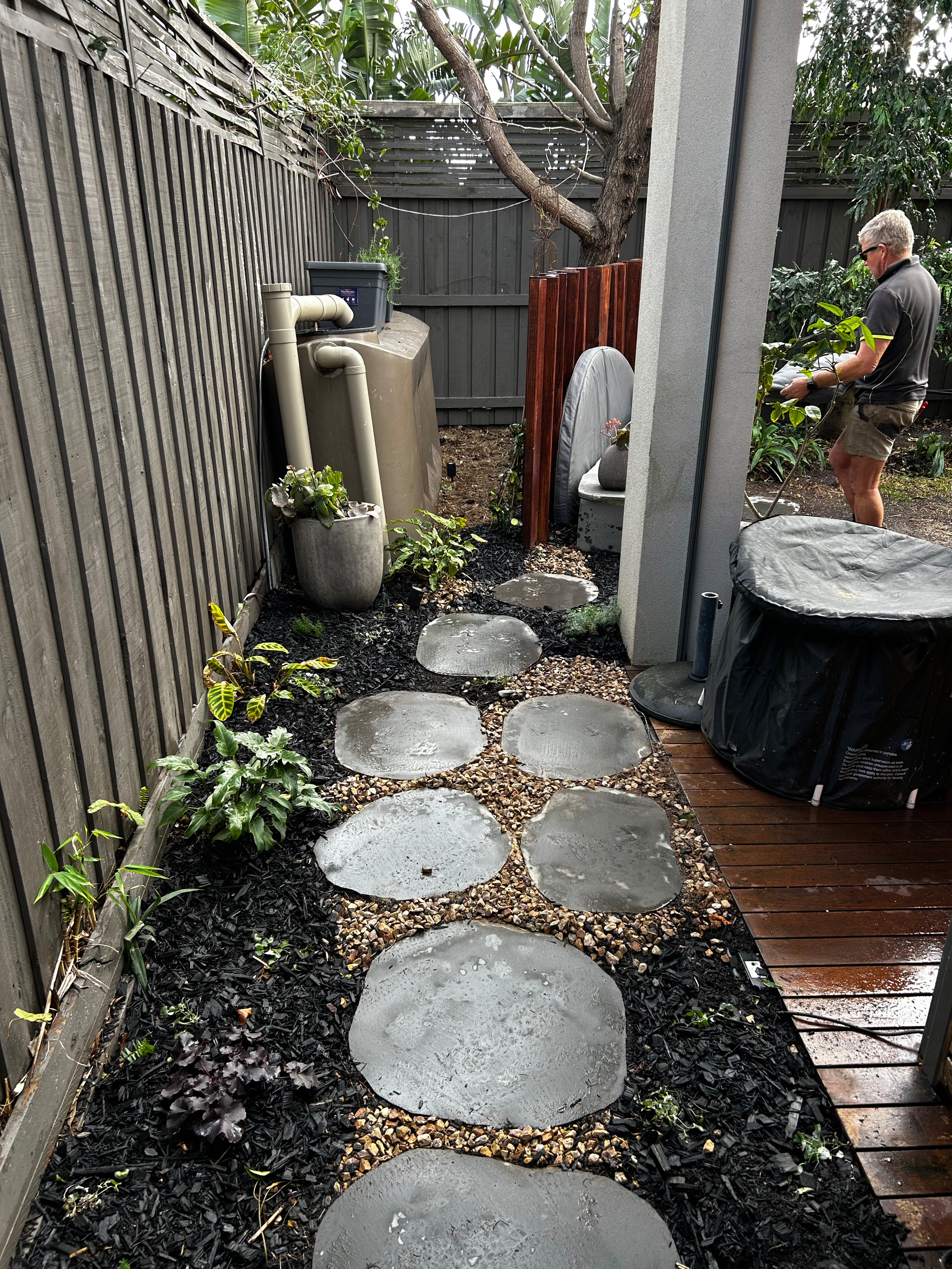 Wet circular stepping stones leading from a wooden deck to a backyard area with a man trimming plants, surrounded by gardening tools, plants, and wooden fencing.