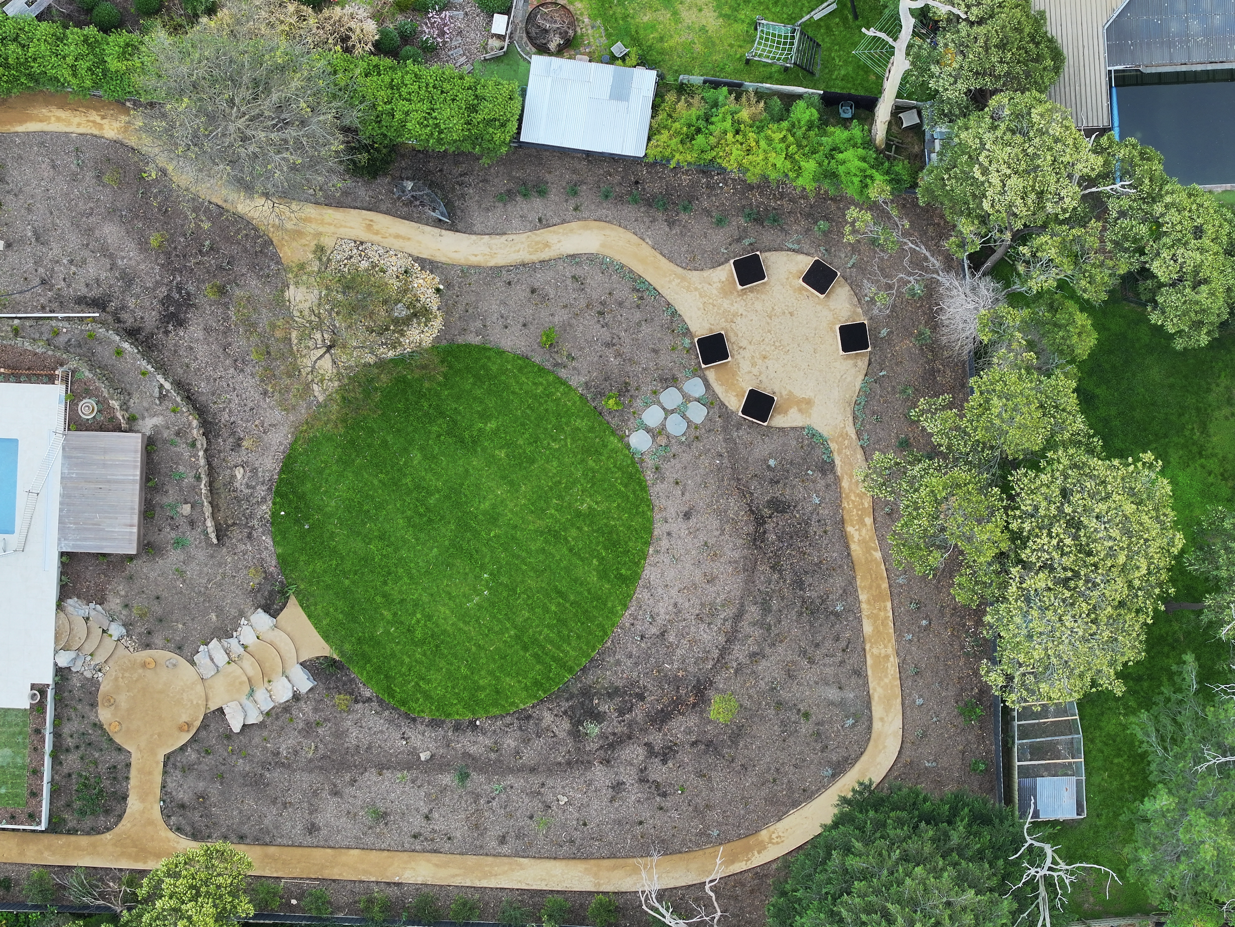An aerial view of a backyard with a circular green lawn, multiple winding dirt paths, and trees. There is a small patio with chairs and tables, a shed, and a structure possibly a greenhouse or small pool. The yard is surrounded by trees and other lan