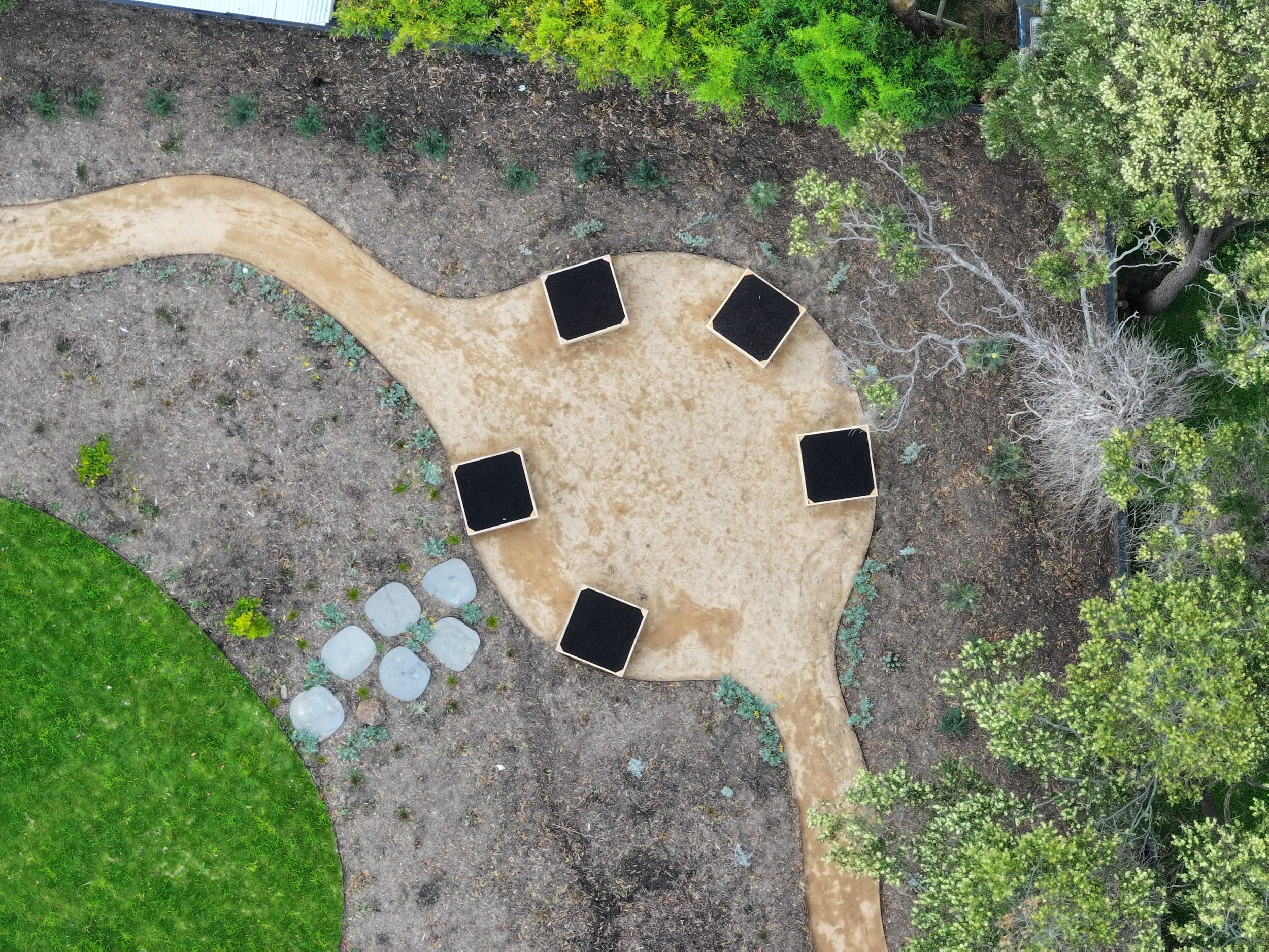An aerial view of a park area with a winding pathway, six black benches arranged around a circular dirt space, a grassy area in the bottom left corner, and surrounded by trees and plants.
