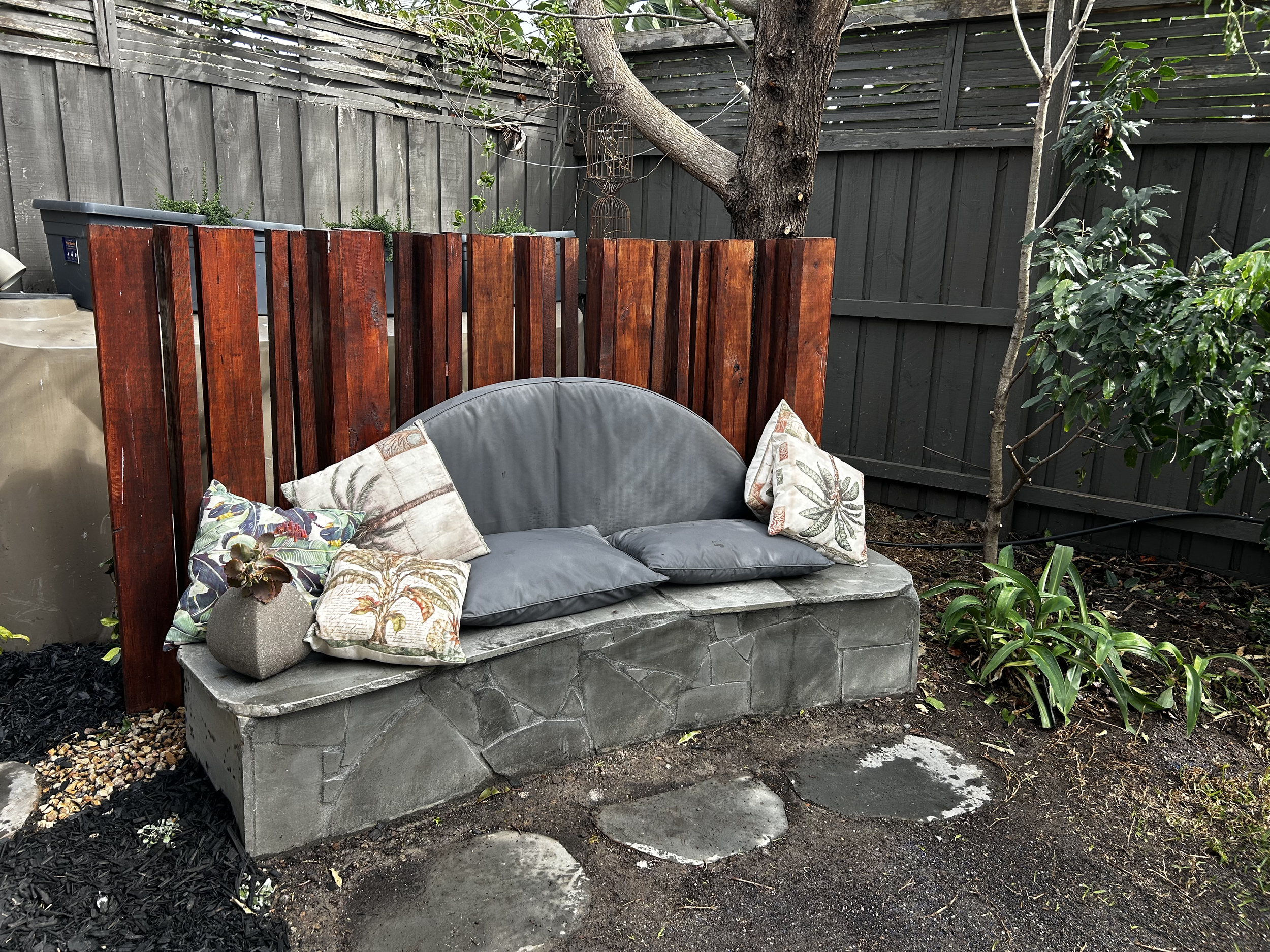 A small outdoor seating area with a gray concrete bench, topped with four decorative pillows and a gray cushion, against a dark wooden privacy fence. A redwood panel extends behind the bench, with a small tree and green plants nearby.