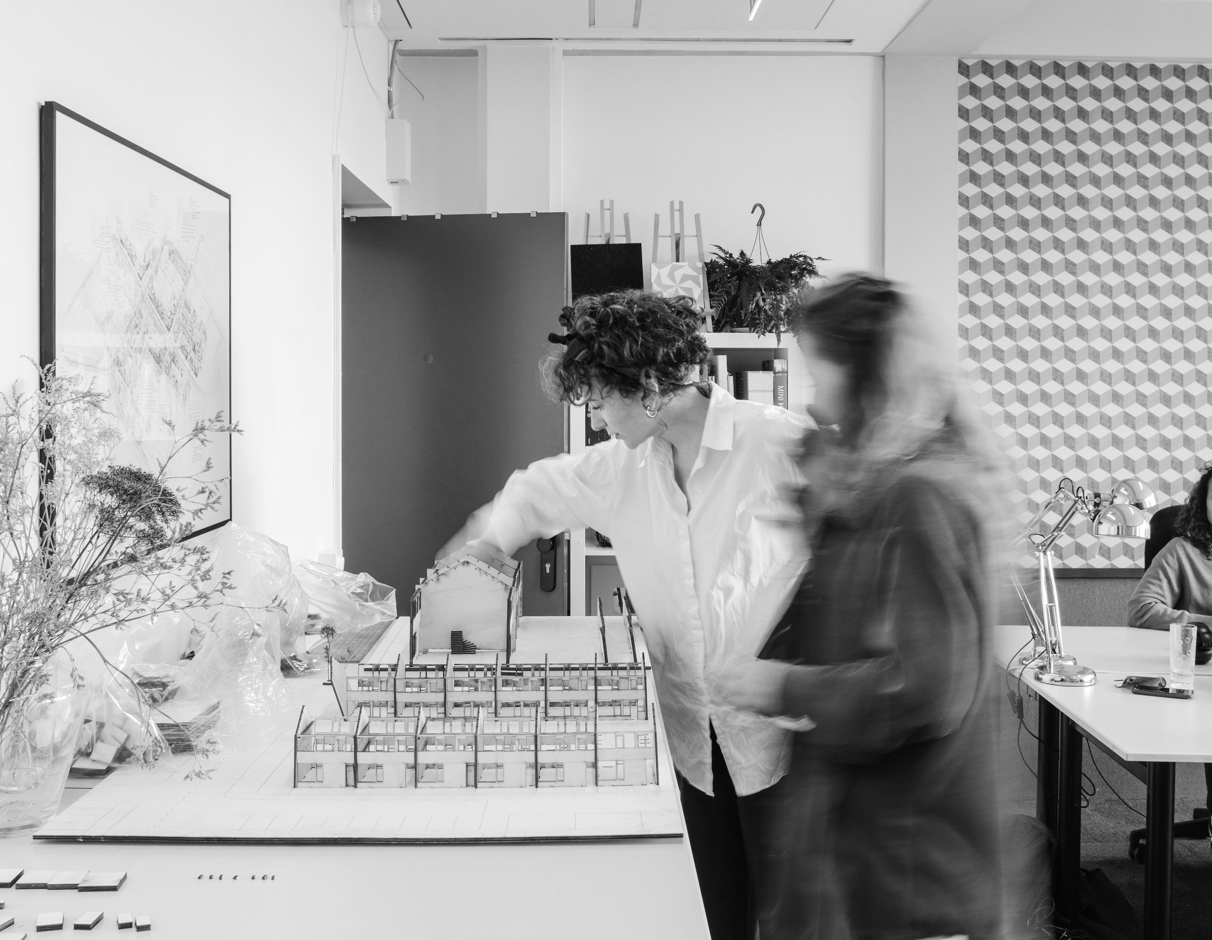 Twee vrouwen bekijken een maquette van een gebouw op een tafel in een witte kamer, op de achtergrond een boekenkast en muur met geometrisch patroon.