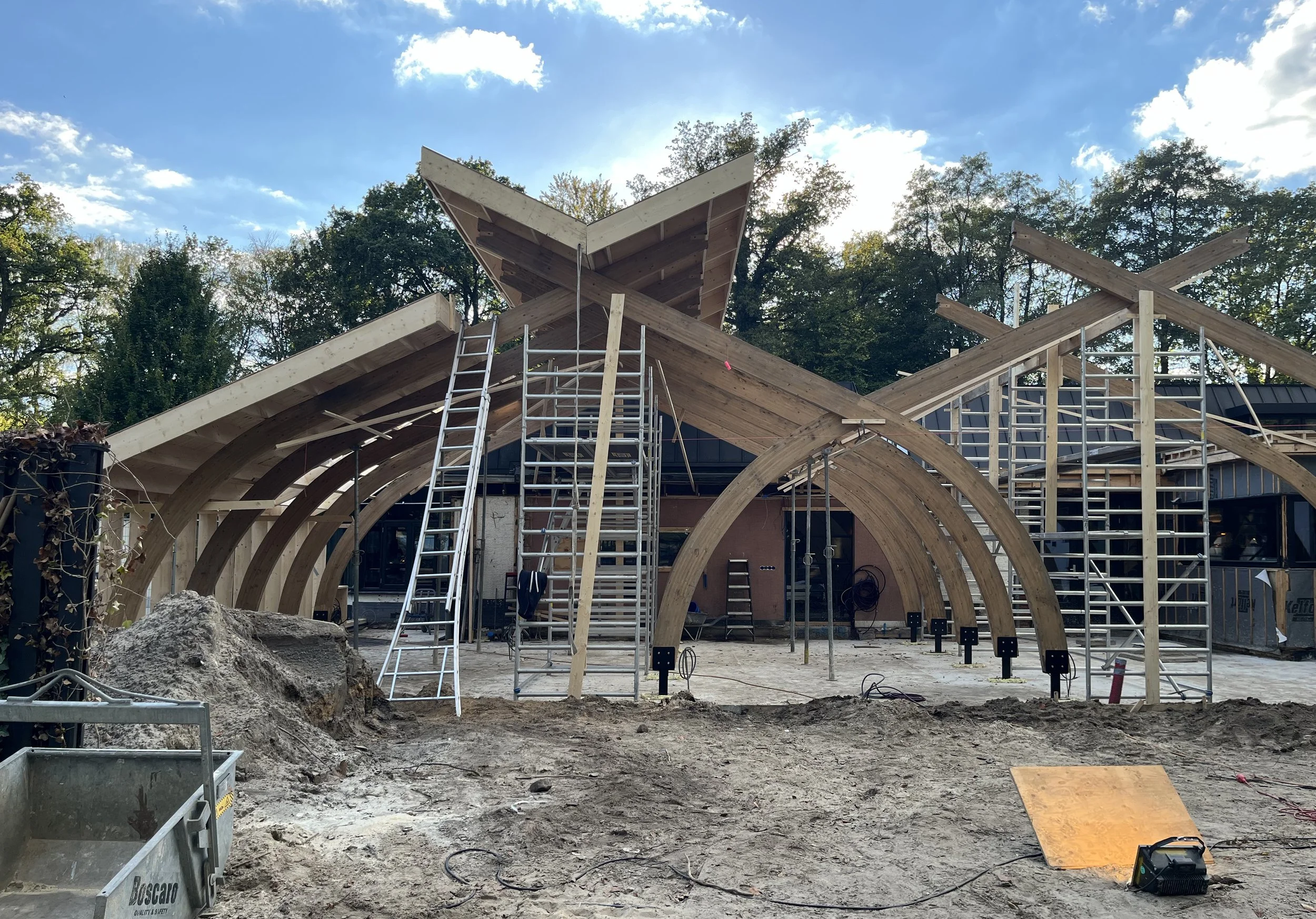 Bouwplaats met houten constructie van hergebruikt hout voor een gebouw, voorzien van ladders en steigers, op een terrein met aarde en bouwmaterialen, temidden van bomen onder een blauwe hemel.