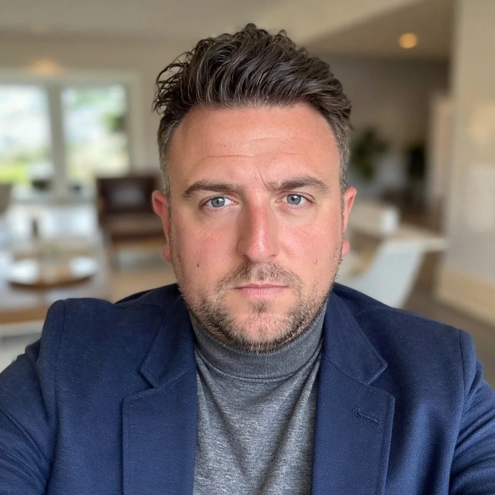 A close-up of a man with styled dark hair, blue eyes, and a beard, wearing a grey turtleneck and blue blazer, with a blurred modern living room background.