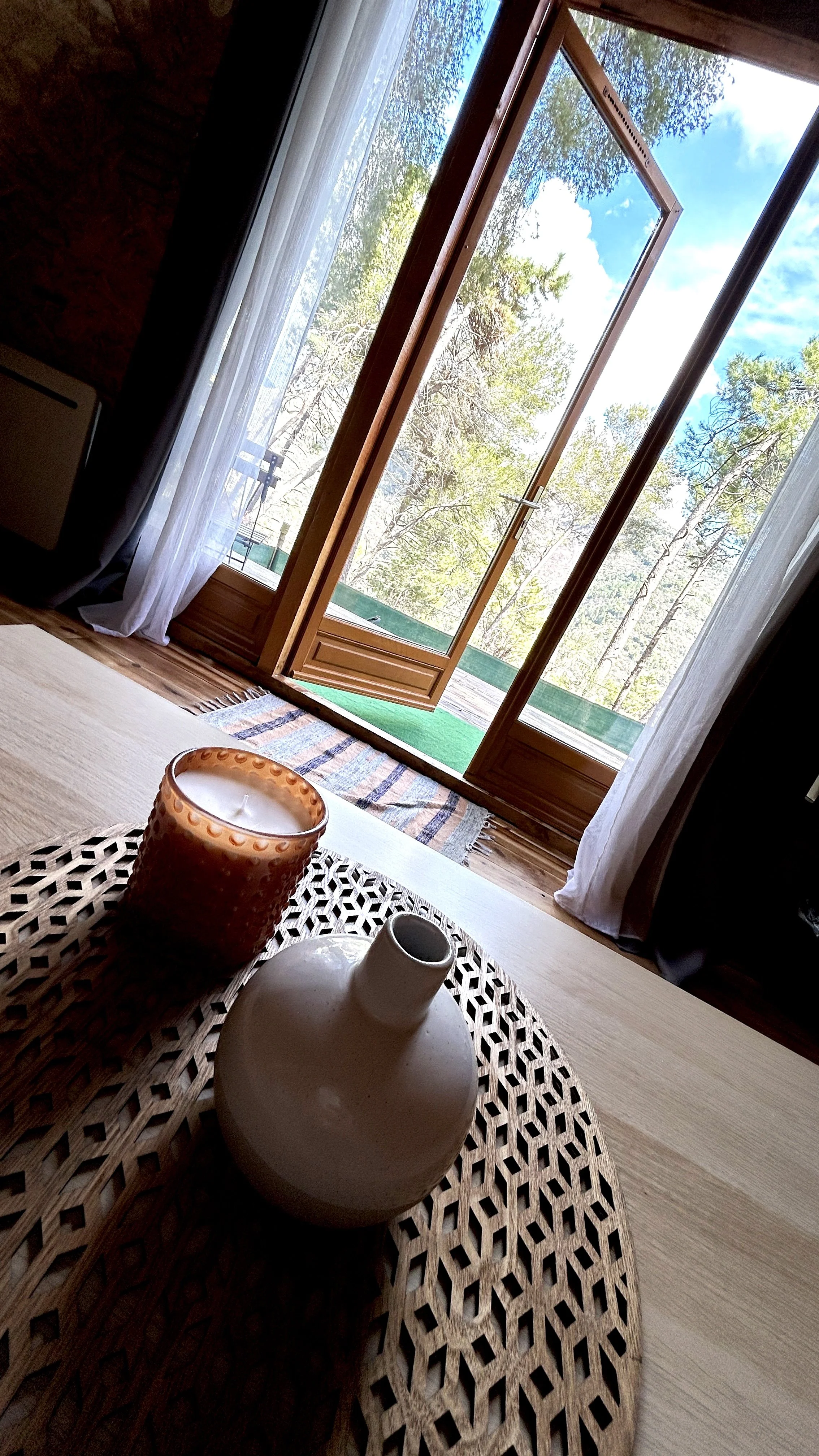 Vue d'une porte-fenêtre ouverte donnant sur une terrasse avec des arbres et un ciel bleu, vue d'un intérieur avec une table en bois, une bougie dans un verre et une petite vasque blanche.