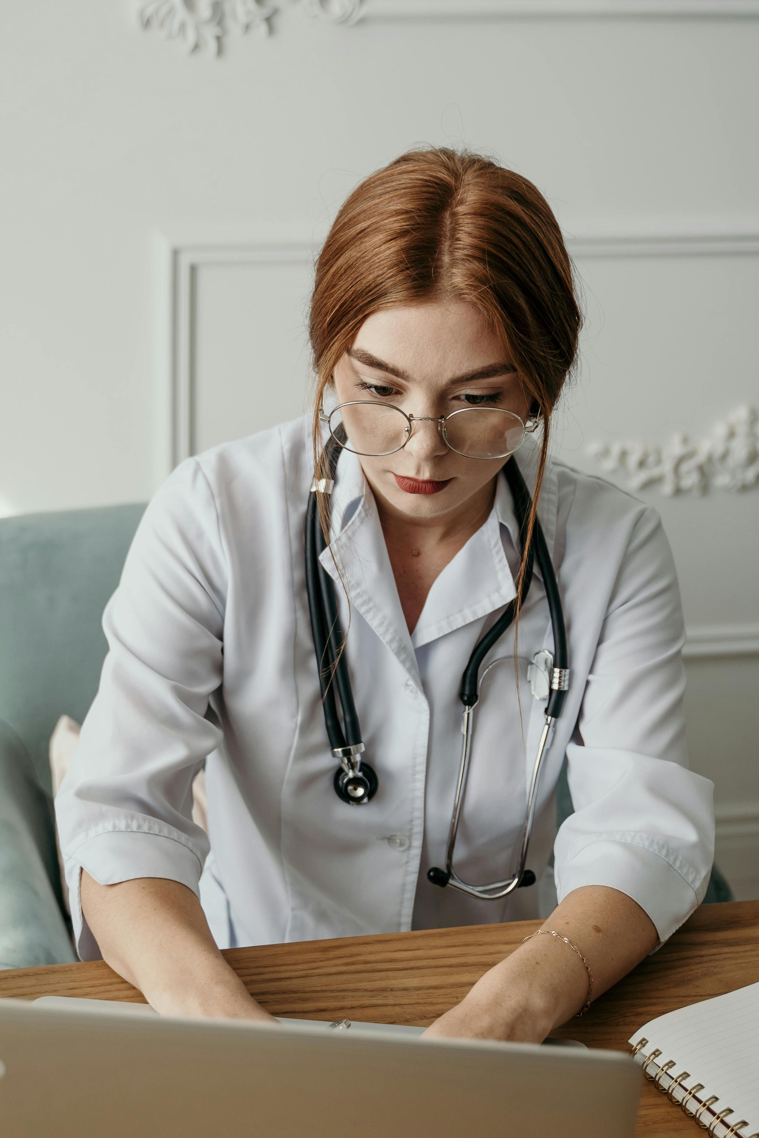 A female healthcare professional or doctor with glasses and a stethoscope around her neck, working on a laptop at a desk.