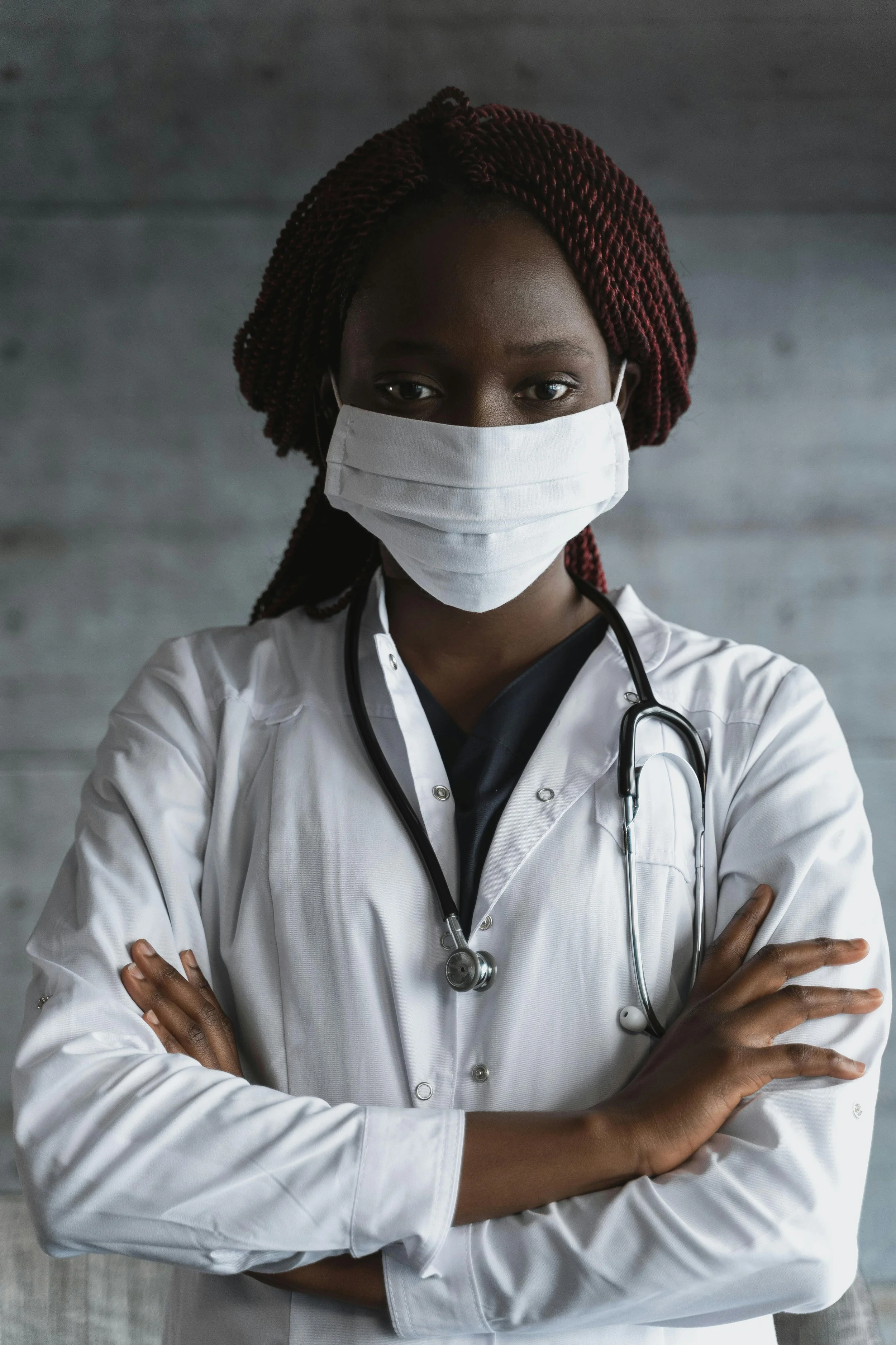 A female healthcare professional wearing a white lab coat, a face mask, and a stethoscope with arms crossed.