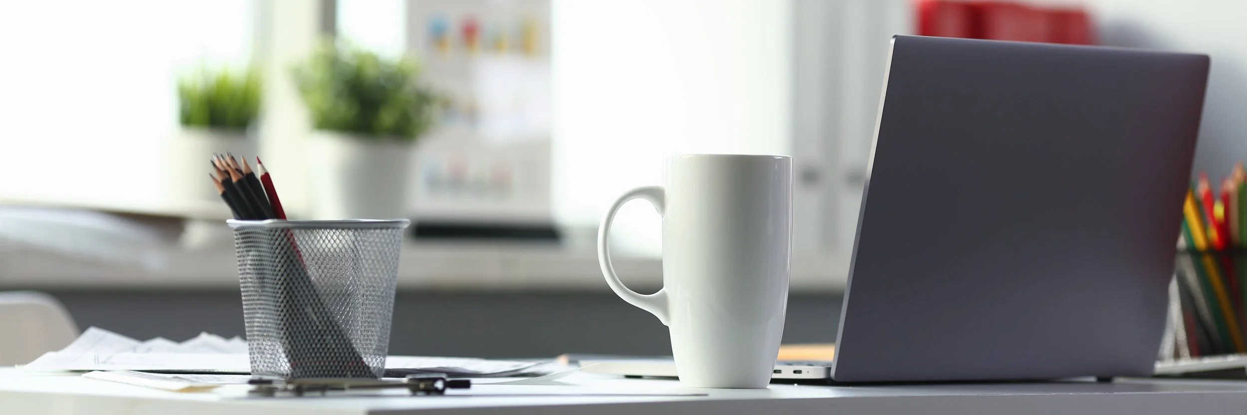 Office desk with a laptop, white coffee mug, container of colored pencils, and scattered papers.