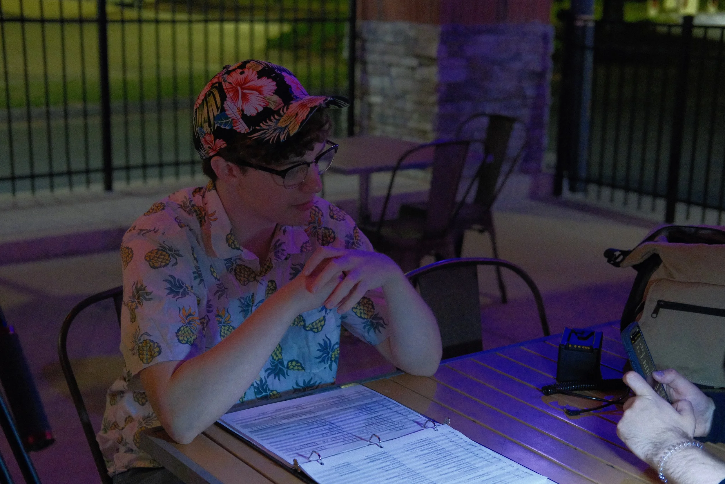 A young man with glasses and a floral Hawaiian shirt, wearing a colorful floral cap, sitting at a table outdoors at night, looking at a menu with someone holding a smartphone nearby.
