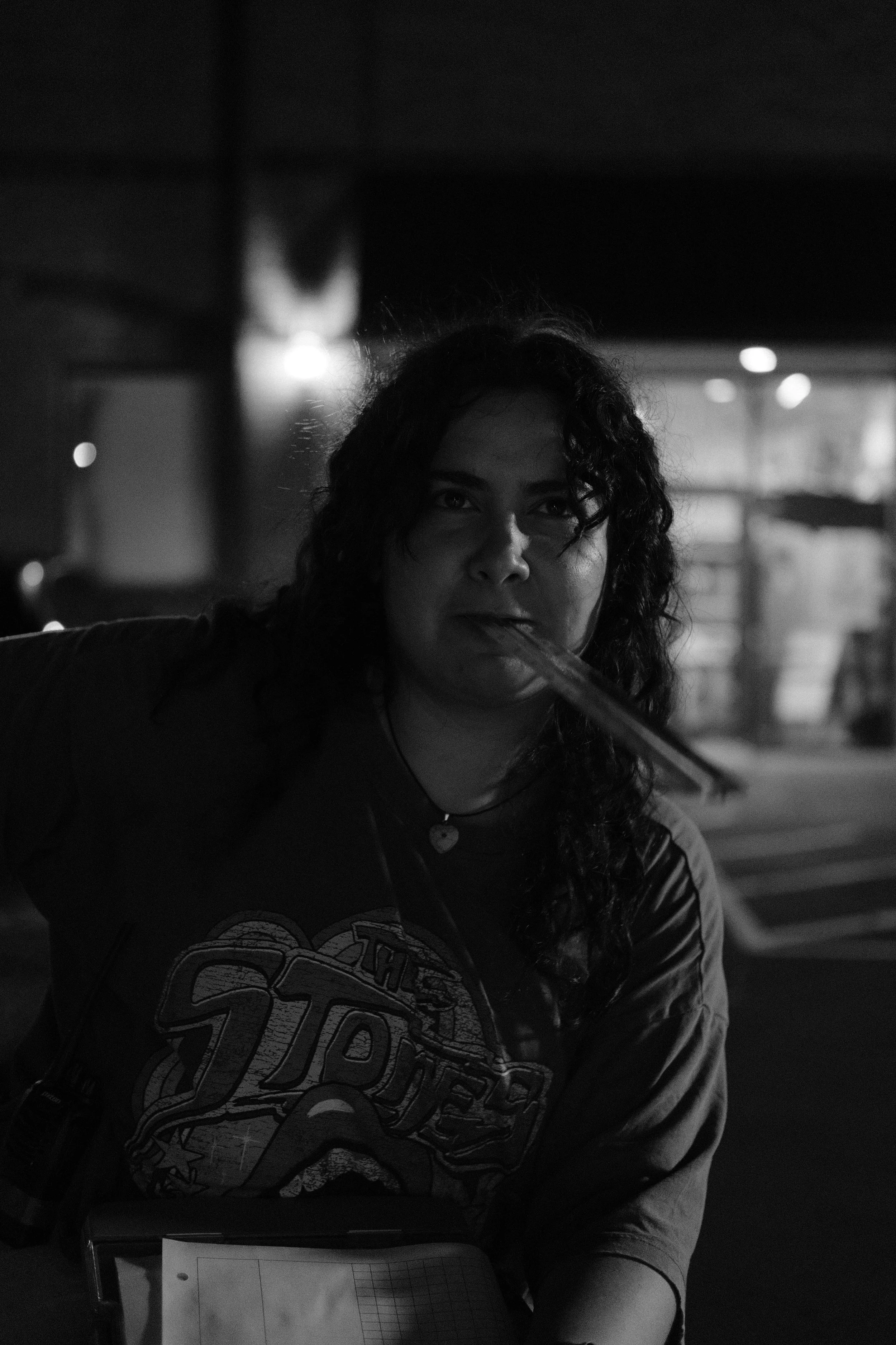 A woman with curly hair standing outdoors at night, holding a pencil in her mouth, wearing a t-shirt with a logo, with lights and a building in the background.