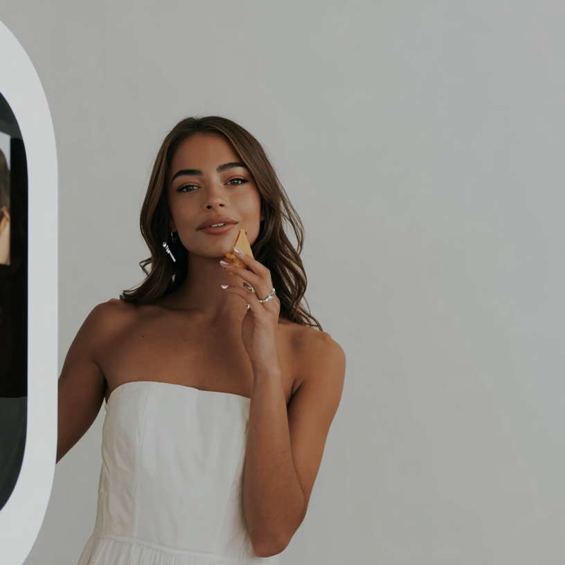 Mlada zena s hnedými vlasy a nausnicemi, oblecena v bilych satatech bez raminek a diva se do kamery. Stoji pred jednolitym svetle sedym pozadim.