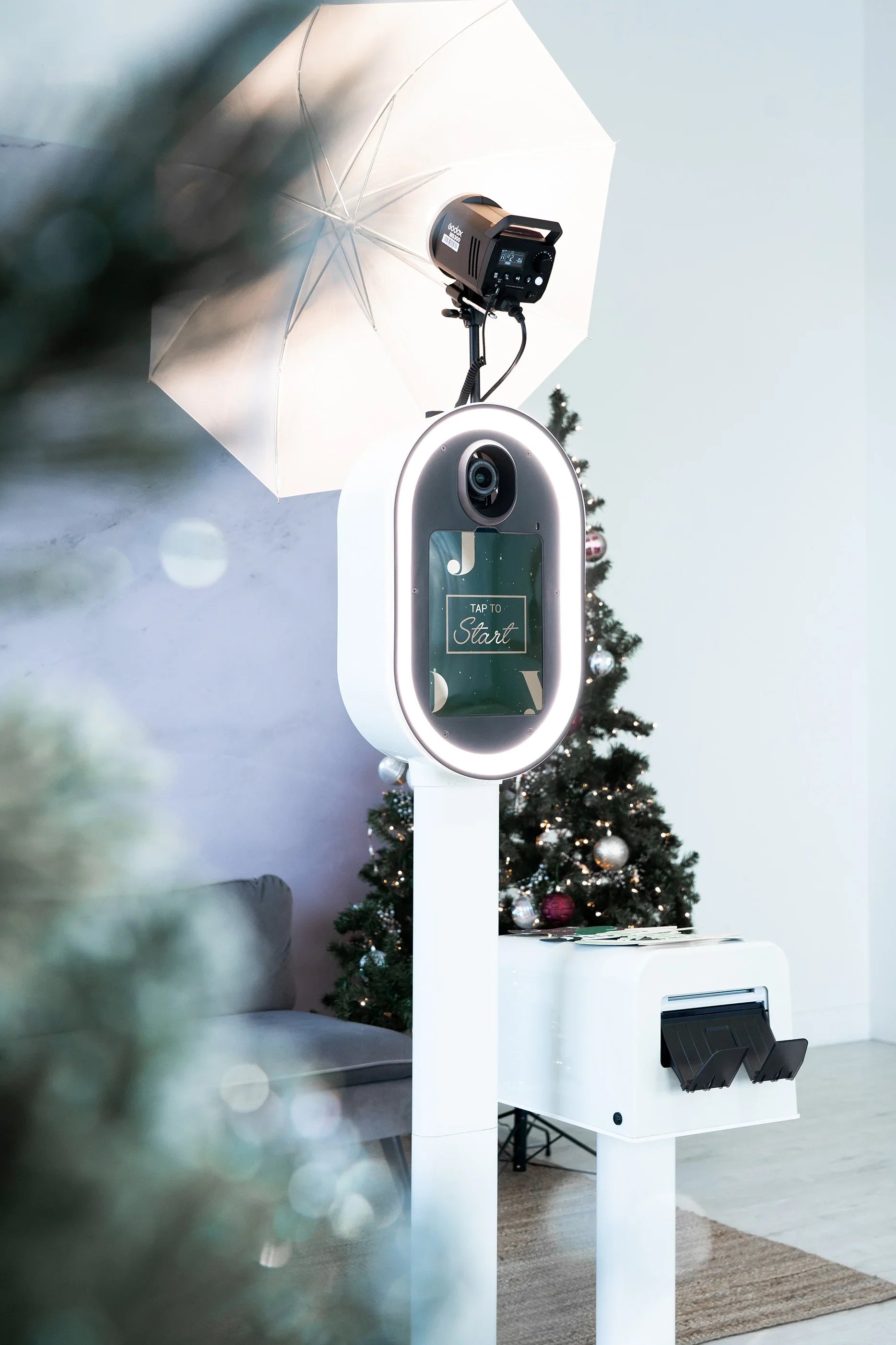 A Christmas tree decorated with ornaments and lights, positioned next to a white photo booth stand with a camera and ring light, and an umbrella light above.
