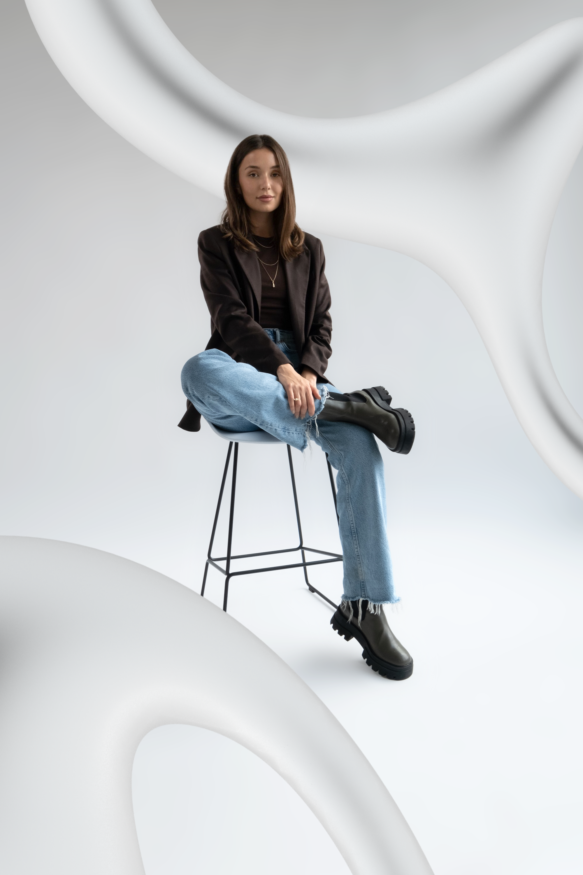 Mlada zena s dlouhymi hnedými vlasy sedi na cerne kovove zidli v minimalistickem studiu a foti se ve fotokoutku