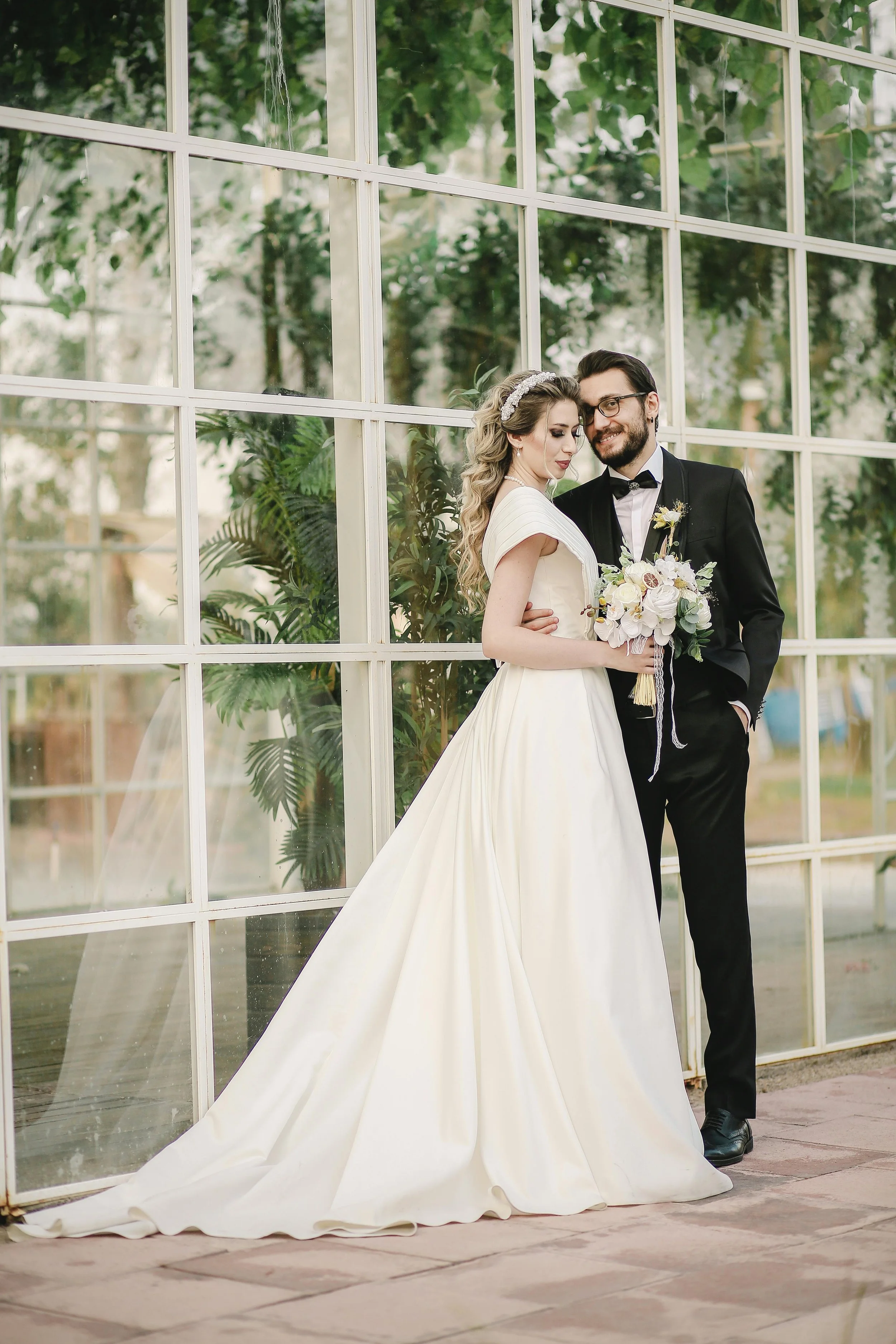 A newlywed couple standing in front of a large glass window, with the bride in a white wedding dress and the groom in a black tuxedo, smiling and holding a bouquet.
